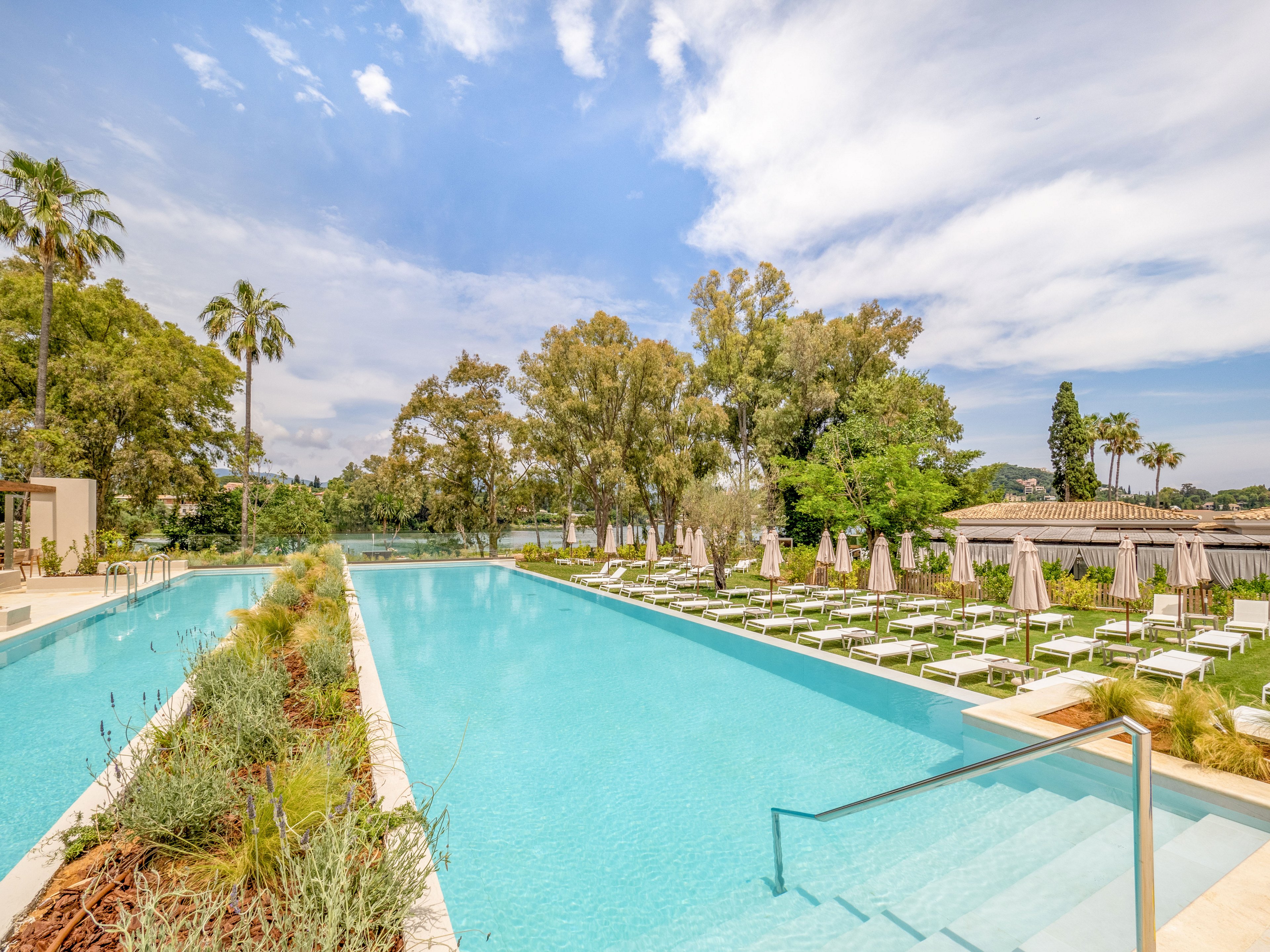 A long, turquoise swimming pool stretches through a landscaped resort surrounded by trees and greenery. White sun loungers and umbrellas line the poolside terrace, creating a relaxing vacation atmosphere. Clear blue sky with scattered clouds adds to the bright, sunny setting. The scene suggests a tranquil, upscale outdoor leisure area ideal for relaxation and sunbathing.