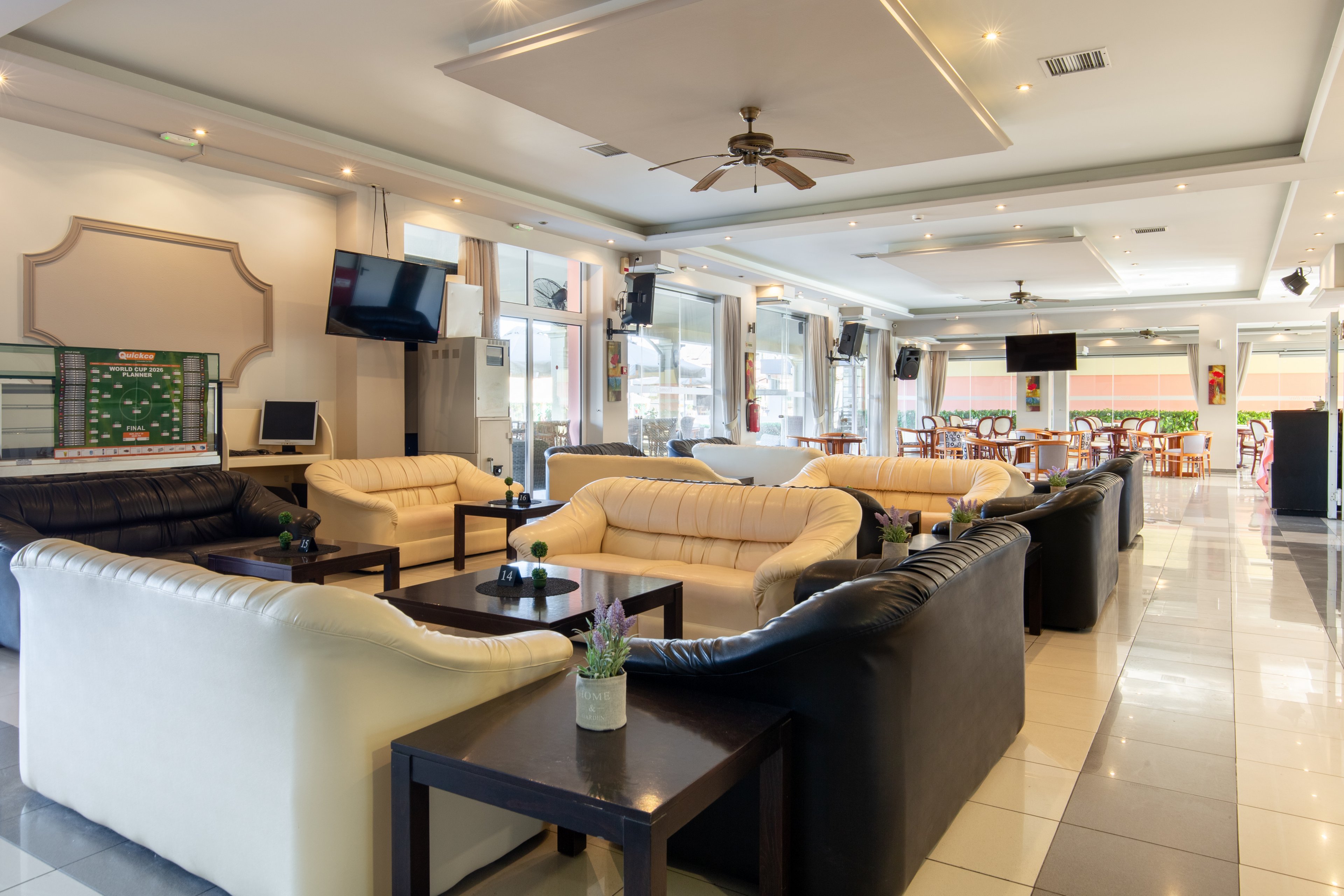A spacious lounge area features cream and dark leather sofas arranged around low wooden coffee tables. The setting appears to be a hotel or resort lobby with tiled floors and large windows letting in natural light. Wall-mounted televisions, ceiling fans, and decorative plants add comfort and visual interest. In the background, additional seating and tables suggest a connected bar or café space.