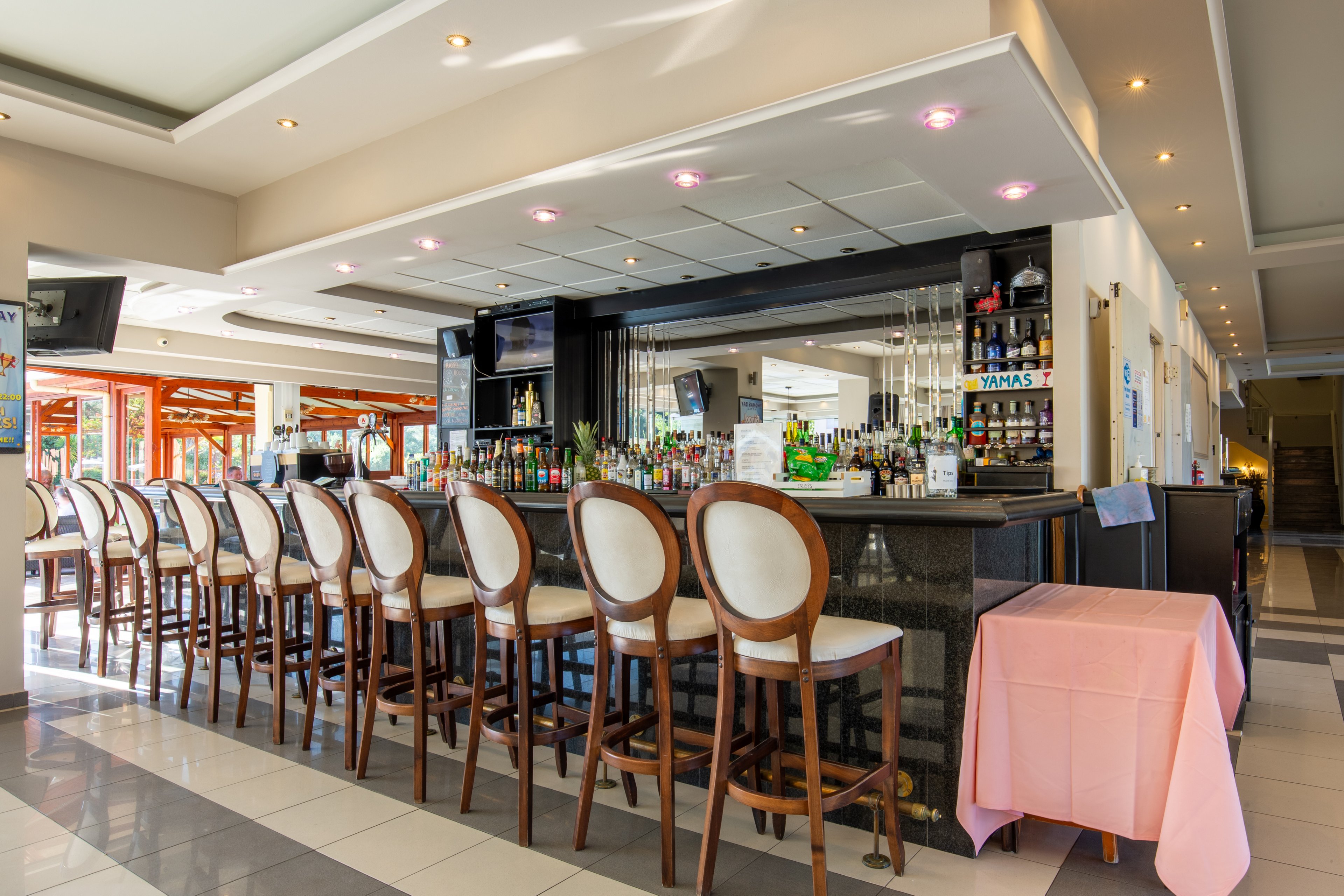 A long row of wooden bar stools with white cushioned backs lines a polished bar counter in a spacious hotel lounge. The bar is stocked with various bottles and glassware, with mirrors and shelves behind the counter. Recessed ceiling lights and large windows create a bright, inviting atmosphere. A small side table covered with a pink tablecloth stands near the bar, adding a soft accent to the modern interior.