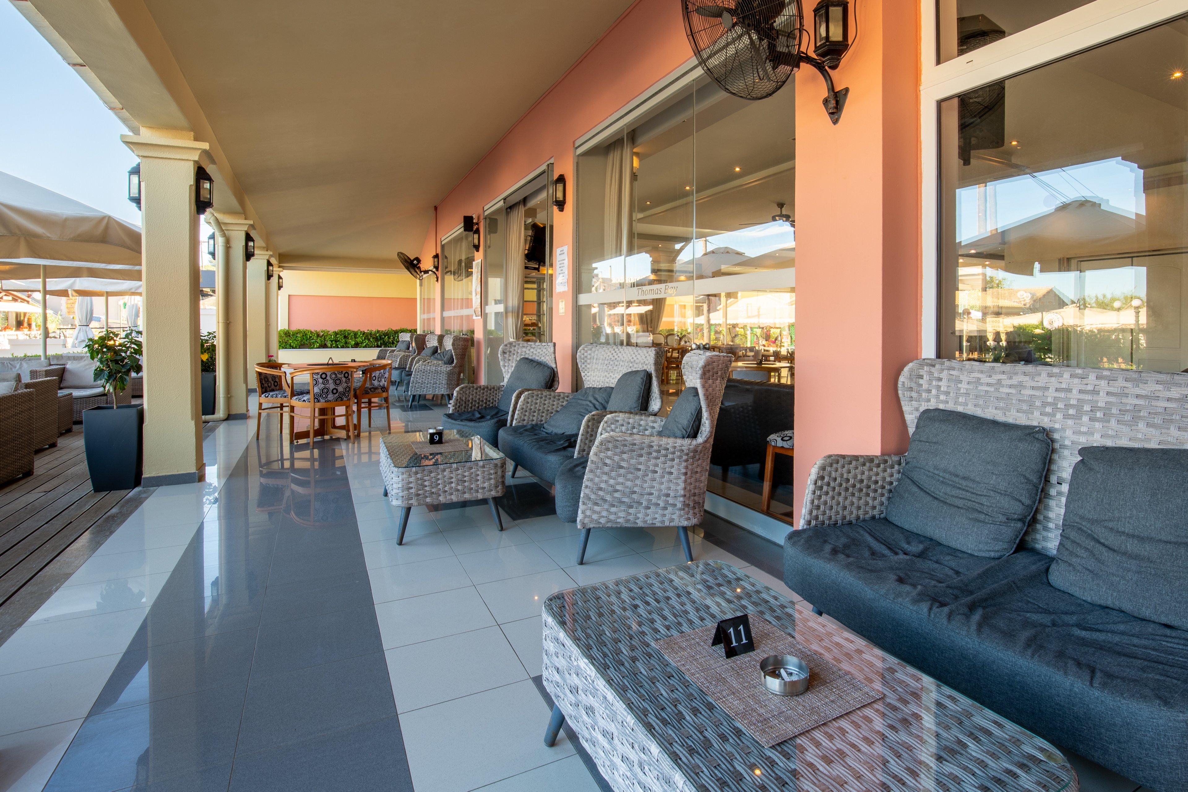 The image shows a spacious outdoor lounge area with wicker sofas and armchairs arranged along a covered terrace. Cushioned seating in neutral gray tones faces small coffee tables, one displaying a table number sign. Large windows and soft lighting reflect the interior of the adjacent building, while umbrellas and additional tables extend the seating area outside. The scene suggests a relaxed resort or restaurant environment designed for comfortable socializing.