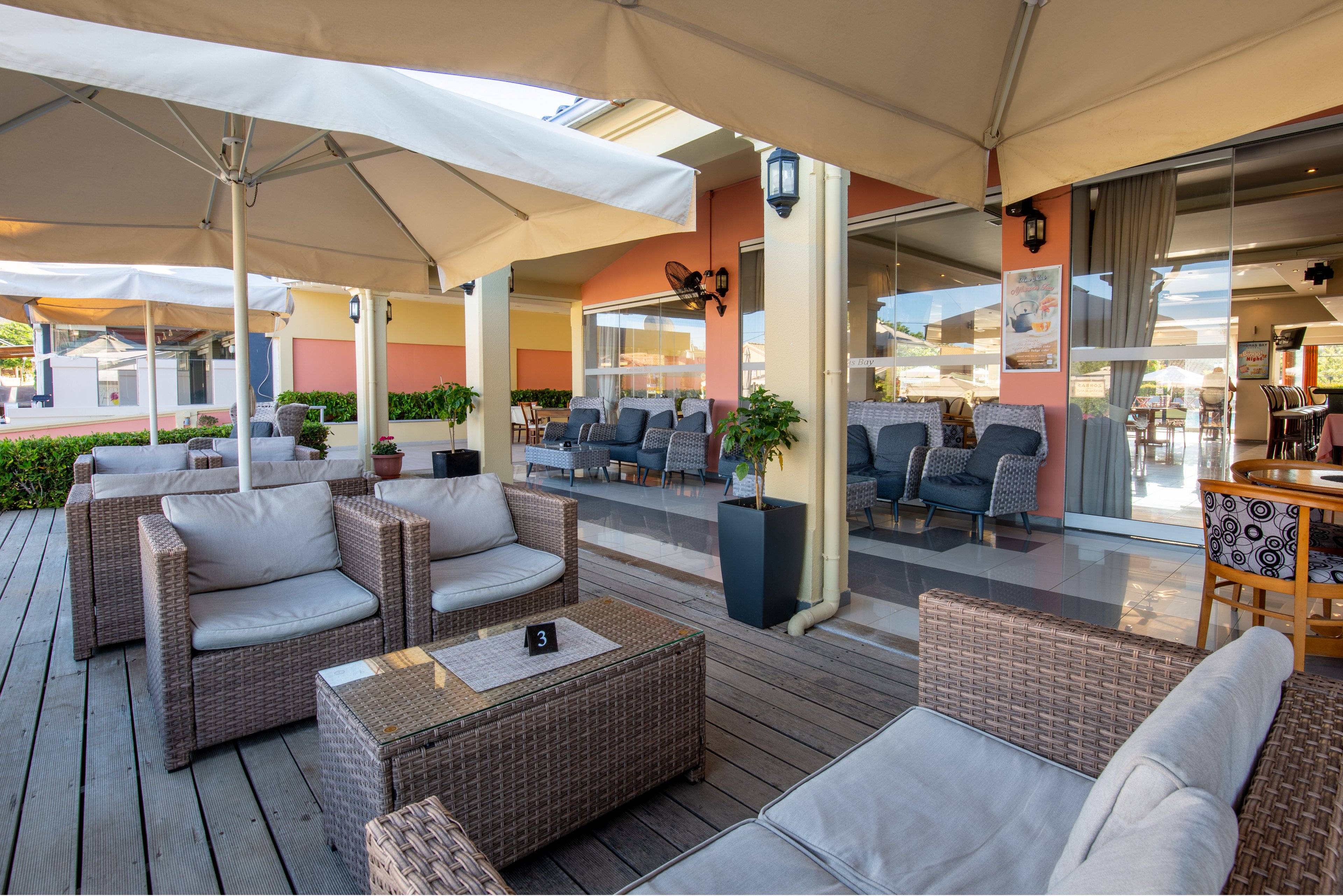 An inviting outdoor lounge area features wicker sofas and armchairs with light cushions arranged on a wooden deck. Large cream-colored canopies provide shade, while potted plants add greenery around the seating. In the background, additional chairs and tables sit near glass doors leading into an indoor space. The scene suggests a relaxed hospitality setting, such as a café, bar, or hotel terrace, on a clear day.