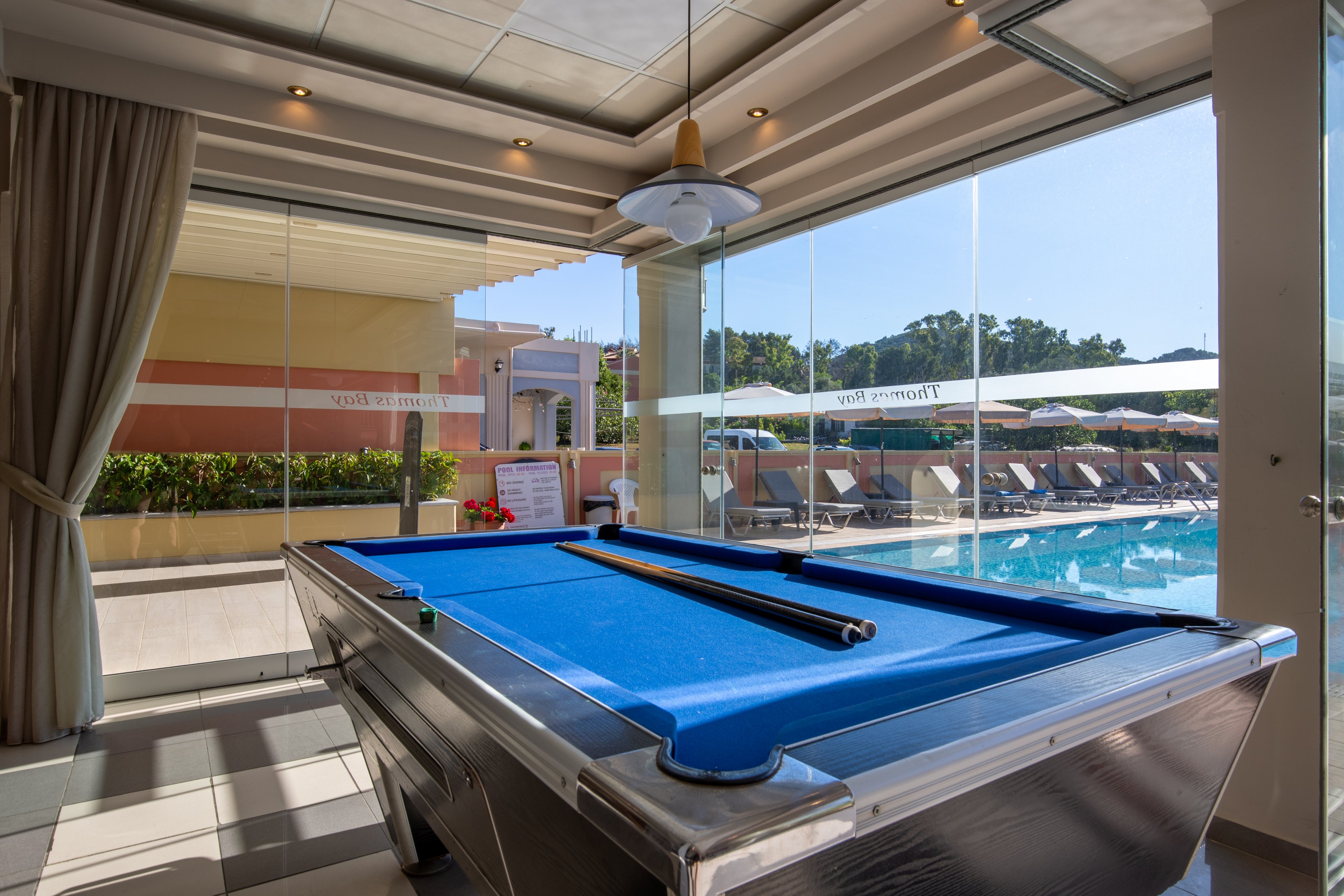 A blue-felt billiards table stands inside a bright room next to large glass windows. Beyond the windows, a sunny outdoor pool area with lounge chairs and umbrellas is visible. Curtains frame the interior space, and a hanging ceiling light is positioned above the table. The scene suggests a relaxed leisure environment combining indoor games with poolside recreation.
