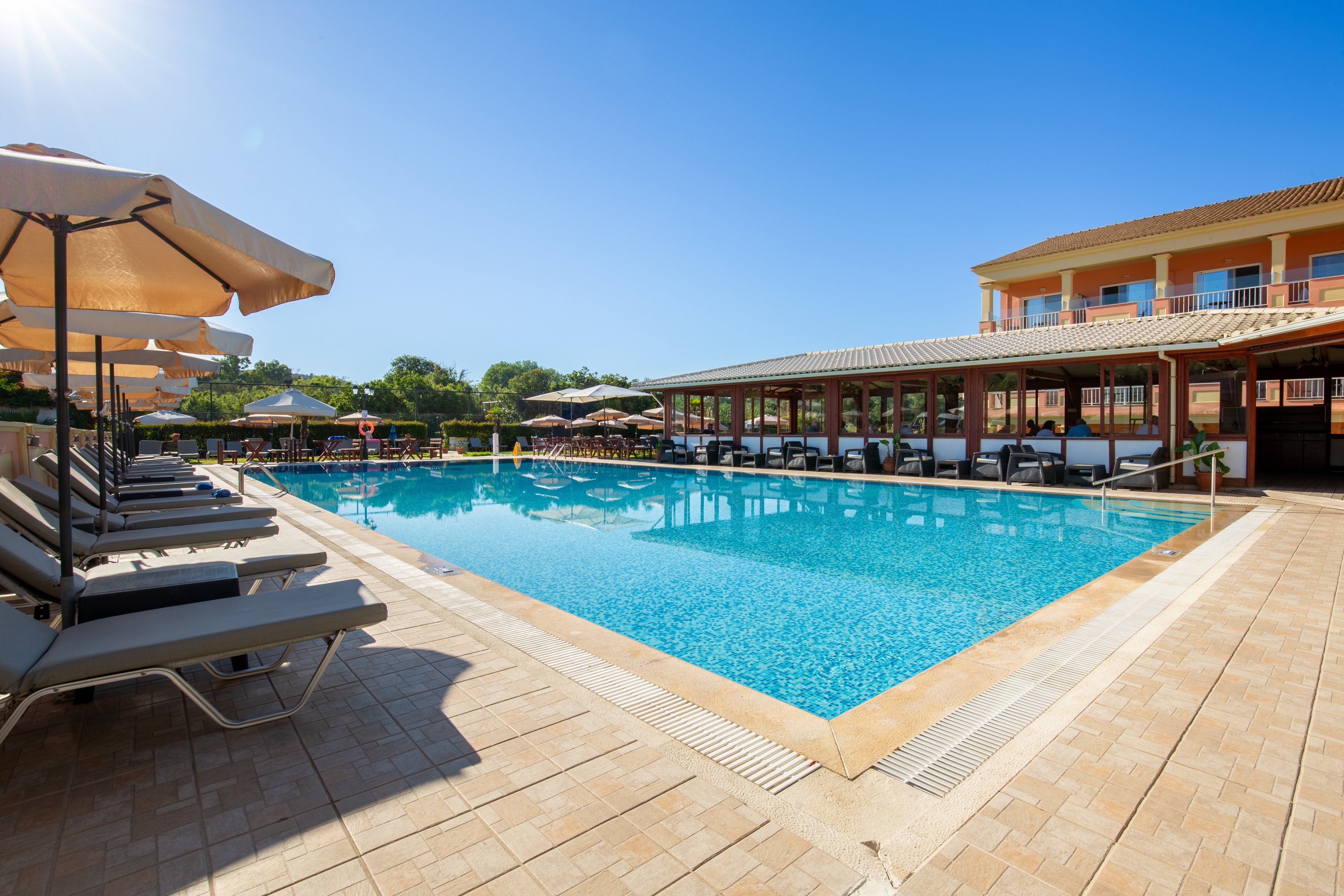 A large outdoor swimming pool is surrounded by sun loungers and umbrellas at a resort. The clear blue water reflects the bright sky, creating a relaxing vacation atmosphere. A multi-story building with balconies and a shaded terrace lines one side of the pool. Trees and greenery are visible in the background, adding to the tranquil holiday setting.