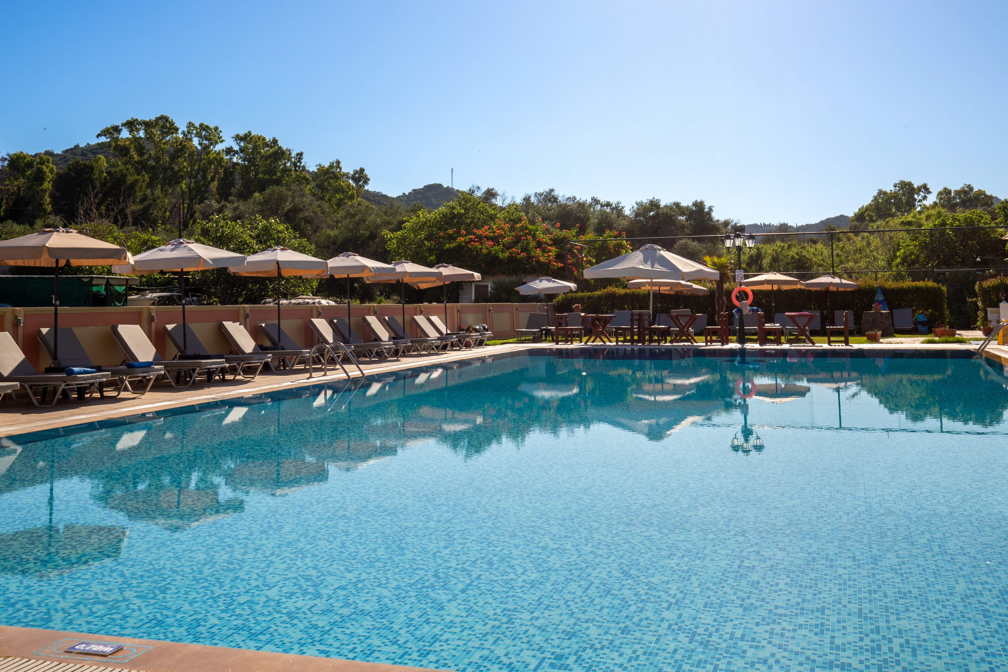 A large outdoor swimming pool glistens under clear blue skies at a resort. Numerous sun loungers and beige umbrellas line the poolside, all currently empty. Green trees, shrubs, and distant hills surround the area, creating a tranquil vacation setting. The calm water reflects the umbrellas and sky, emphasizing the relaxed, peaceful atmosphere.