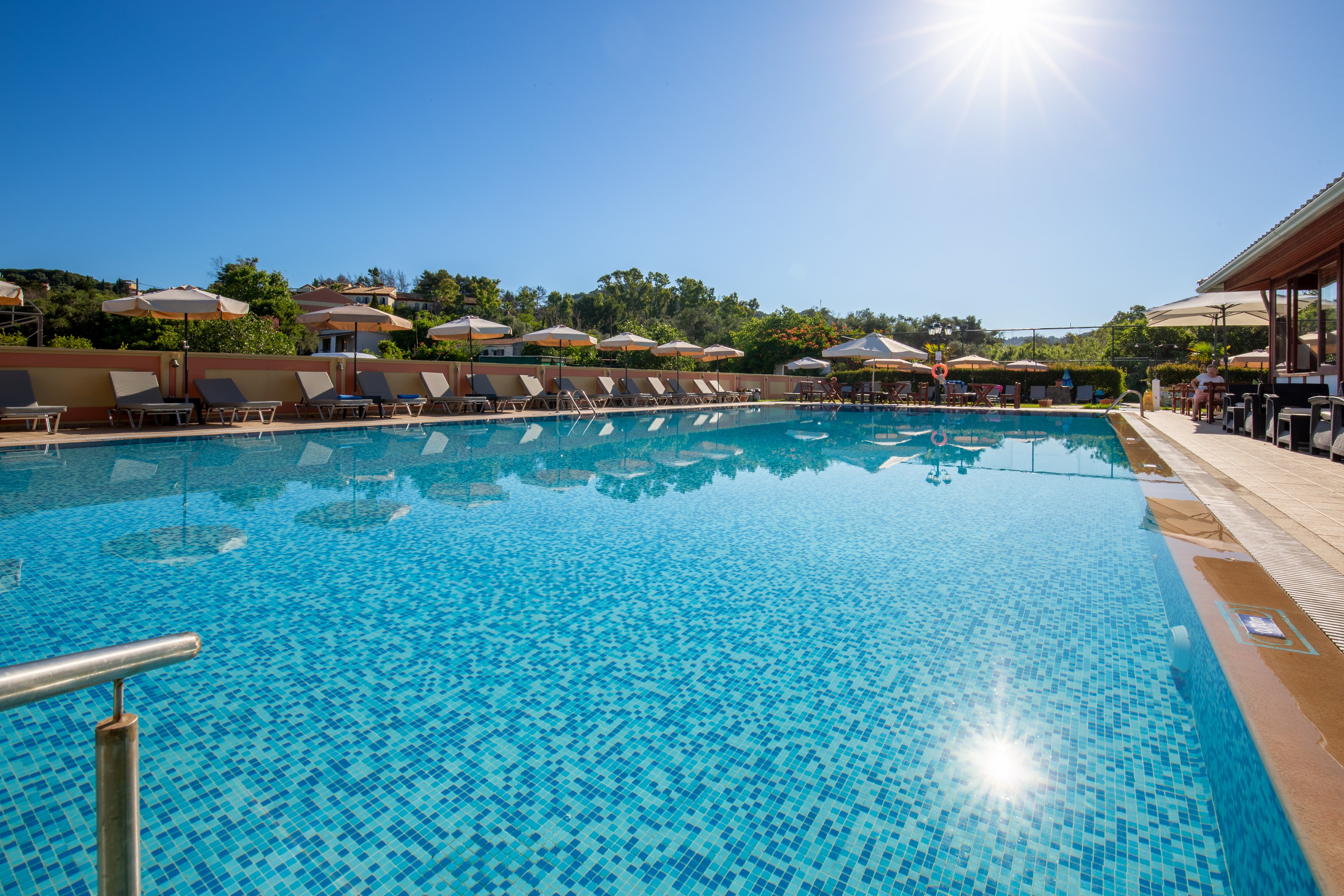 A large outdoor swimming pool glistens under a bright sun in a clear blue sky. Sun loungers and umbrellas line the poolside, suggesting a relaxing resort environment. Trees and greenery form a natural backdrop beyond the pool area. The calm water and warm lighting create a tranquil vacation atmosphere.