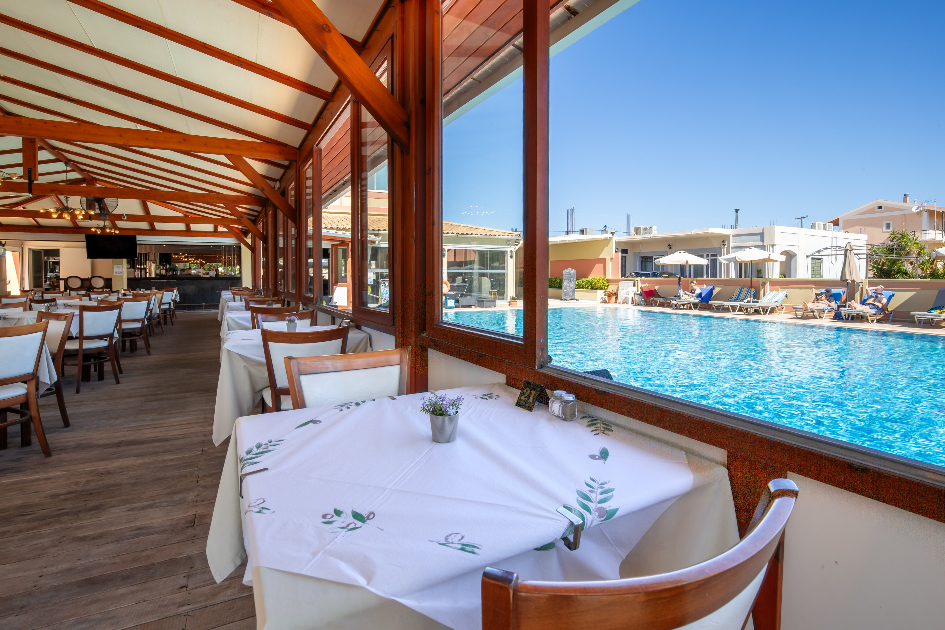 Bright poolside restaurant tables are set with white cloths and small potted plants. Large windows and a wooden ceiling frame the view of a clear blue swimming pool. Sun loungers and umbrellas line the poolside, suggesting a relaxed vacation atmosphere. The scene is empty of people, emphasizing calm, clean, and inviting surroundings.