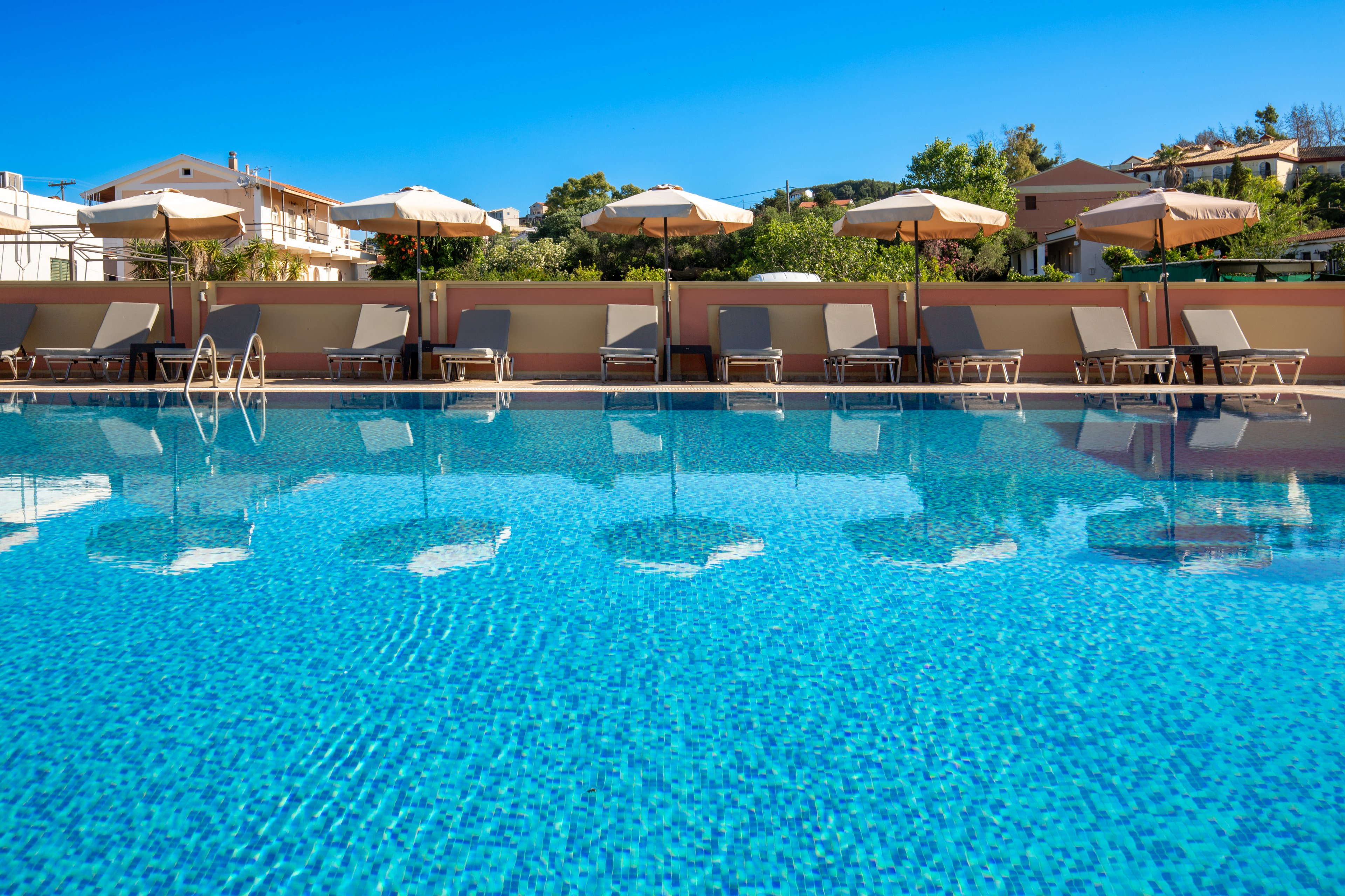 A clear blue outdoor swimming pool reflects sun umbrellas and lounge chairs arranged along the deck. The scene is set at a resort or hotel with low-rise buildings and greenery in the background. Bright sunlight and a cloudless sky create a warm, inviting atmosphere ideal for relaxation. The image emphasizes leisure, vacation, and tranquil summer vibes.