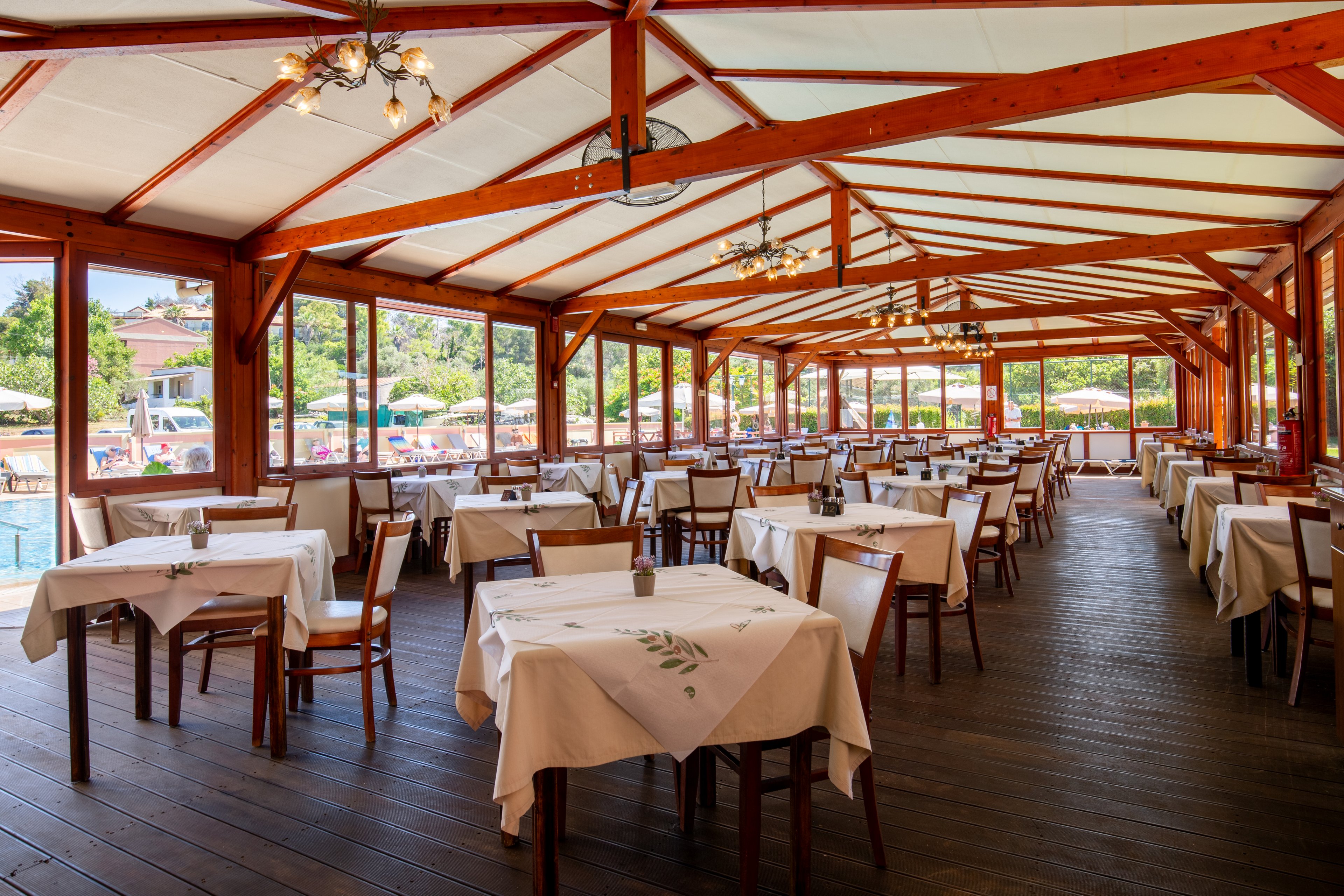 Spacious open-air restaurant dining area features rows of wooden tables and chairs neatly arranged with white tablecloths. Large windows and open sides reveal a sunny poolside setting with umbrellas and greenery outside. Warm wooden beams and ceiling structures create a cozy, rustic atmosphere. The space appears ready for guests, offering a relaxed vacation or resort dining experience.