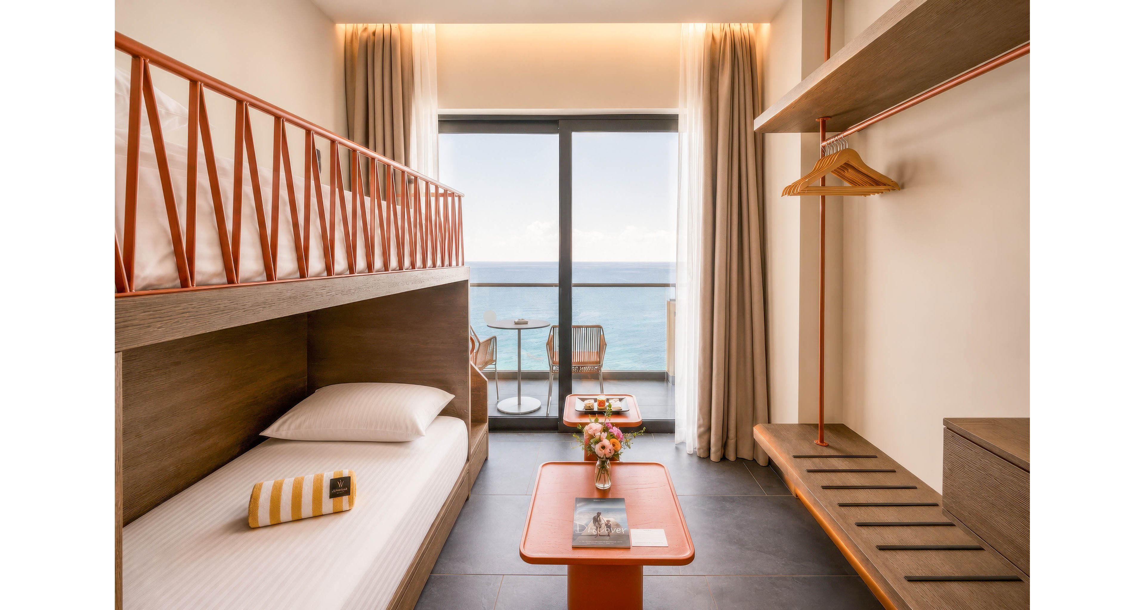 Contemporary hotel room features a wooden bunk bed and minimalist open wardrobe. Large floor-to-ceiling windows open onto a balcony overlooking the ocean with two chairs and a small table. Neutral tones of beige and wood are accented by warm copper details and soft lighting. A striped towel on the bed and fresh flowers on the table add subtle color and a welcoming touch.