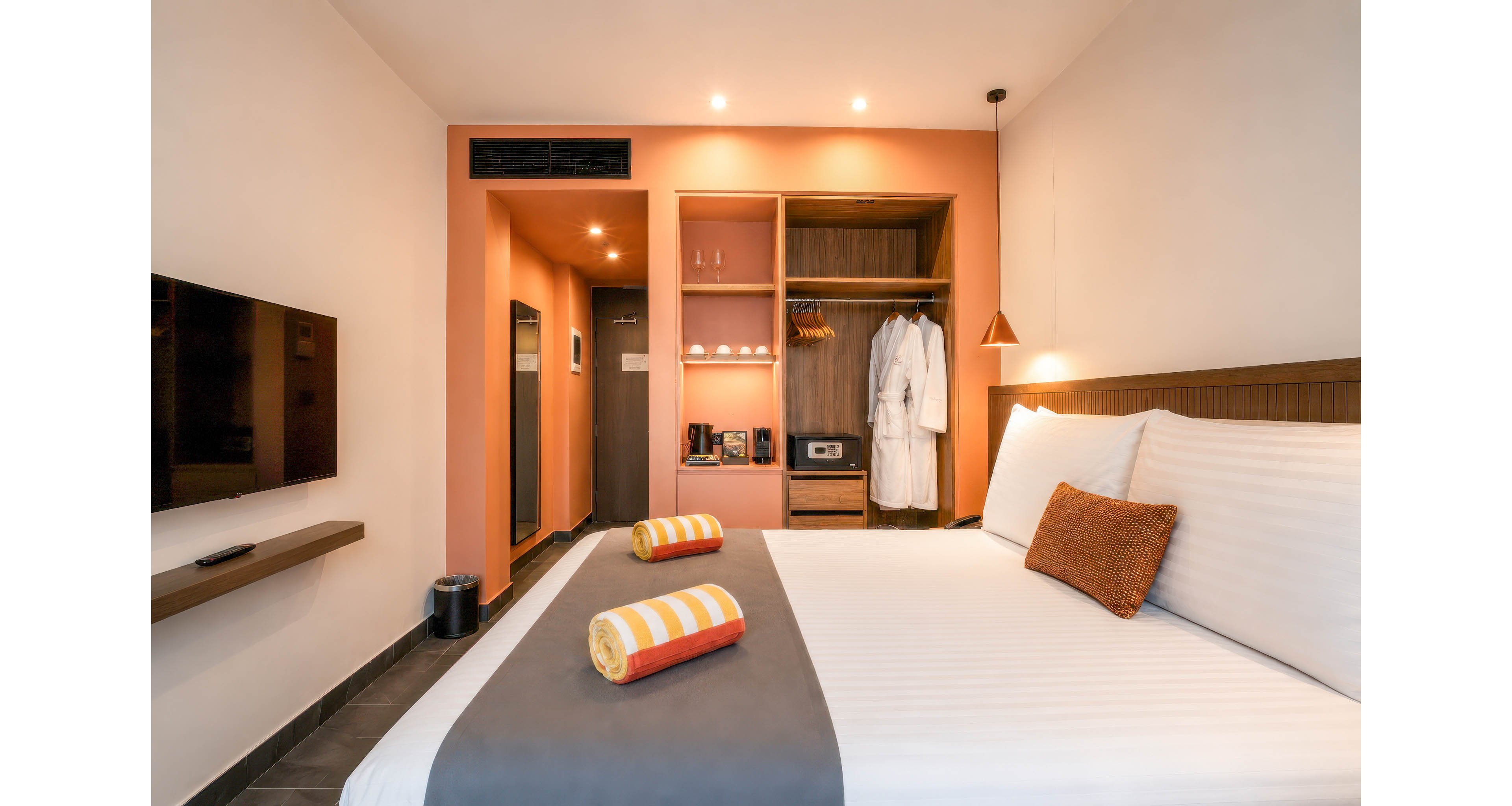 A contemporary hotel room features a neatly made bed with white linens and a gray runner. Two rolled striped towels rest on the bed, adding a pop of color to the neutral decor. An open wardrobe reveals bathrobes, shelves with amenities, and a small safe. Warm recessed lighting and a wall-mounted television create a cozy, inviting atmosphere.