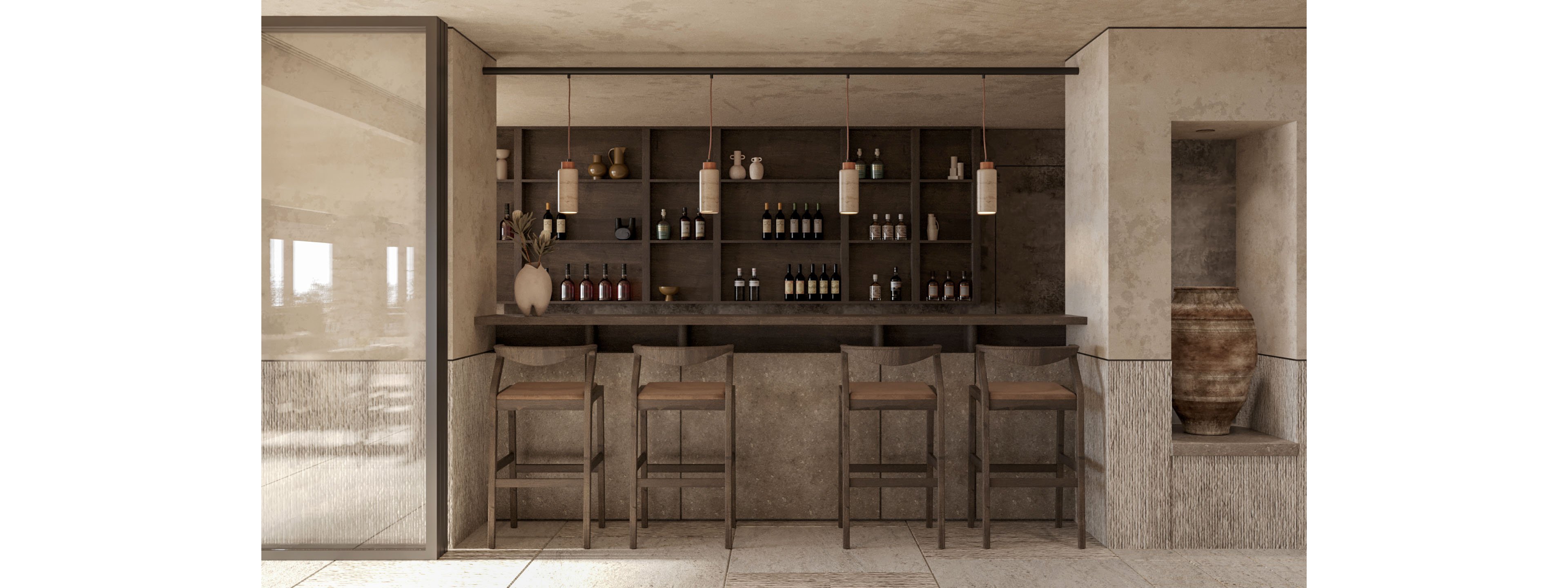 A minimalist bar interior features a long counter with four wooden bar stools arranged in a row. Behind the counter, shelves display an assortment of bottles and glassware, framed by simple vertical panels. Pendant lights with cylindrical shades hang evenly above the bar, casting a warm, soft glow. The space is finished in neutral beige and taupe tones, with textured stone walls and flooring. A large ceramic urn stands in a recessed niche, adding a rustic accent to the contemporary design.