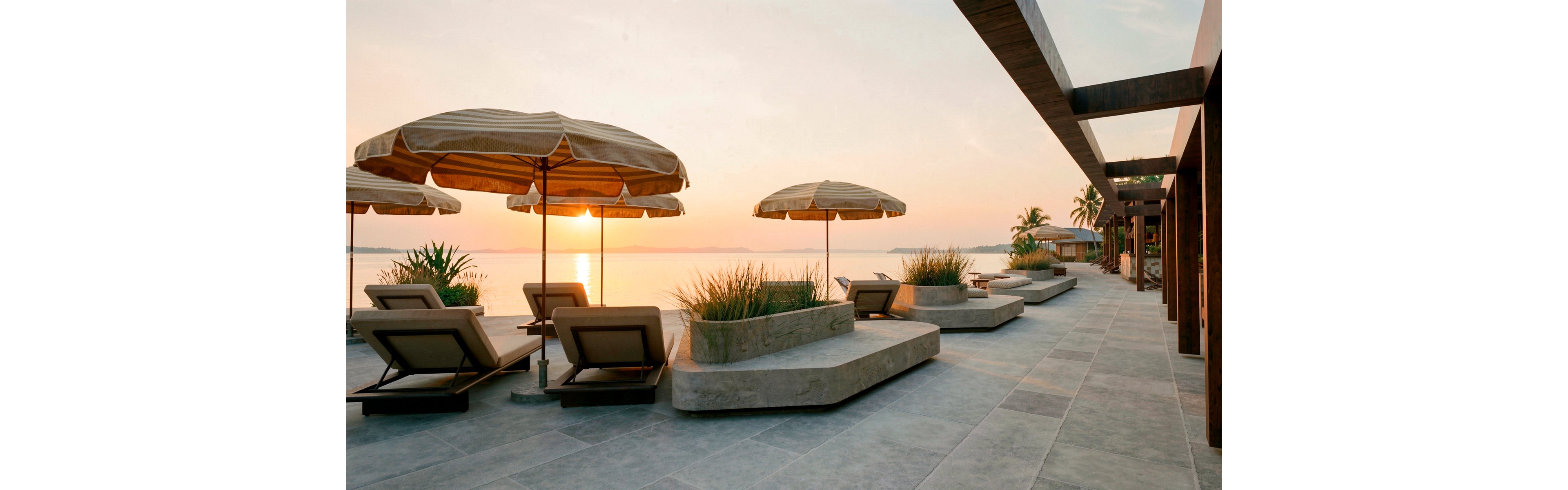 A modern beachfront terrace features cushioned sun loungers arranged beneath large parasols facing the water. The scene overlooks a calm sea at sunset, with warm light reflecting on the expansive stone-tiled floor. Minimalist concrete planters with ornamental grasses add texture and greenery to the contemporary design. Palm trees and distant shoreline elements enhance the relaxed, resort-style atmosphere.