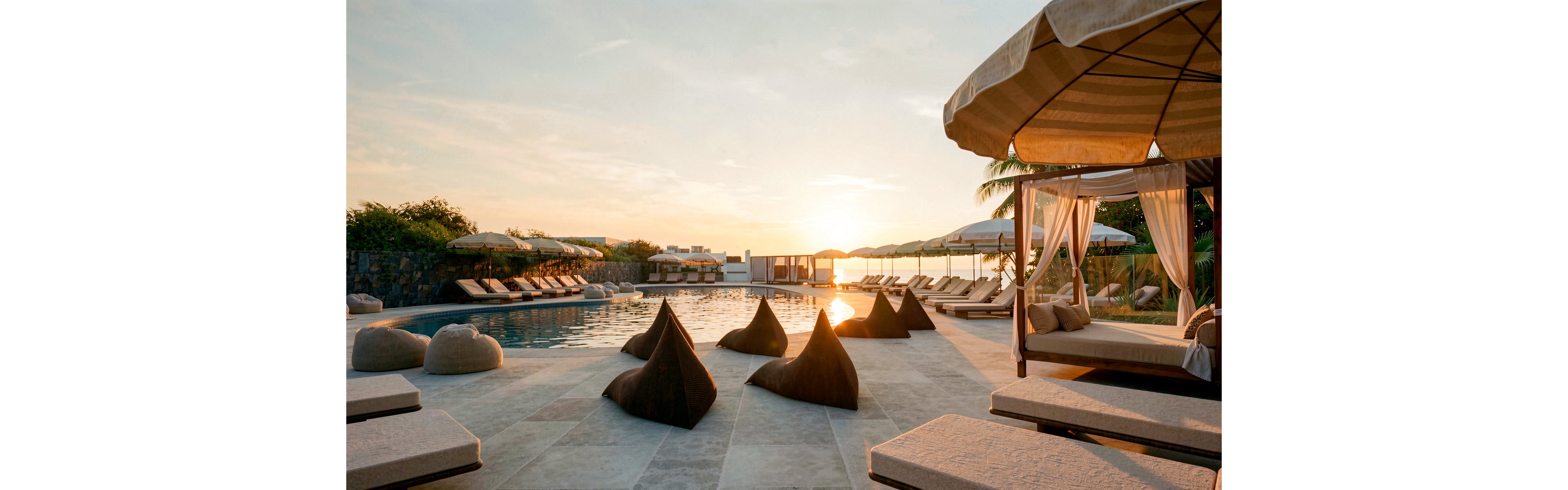 An upscale resort pool area is shown at sunset, featuring lounge chairs and shaded cabanas arranged around a tranquil swimming pool. Soft beanbag-style seats and cushioned sunbeds line the stone-tiled deck, creating a relaxed vacation atmosphere. Large umbrellas and a canopy bed frame the scene, while palm trees and distant greenery suggest a tropical or coastal location. Warm golden light reflects off the water, emphasizing a serene and inviting mood.
