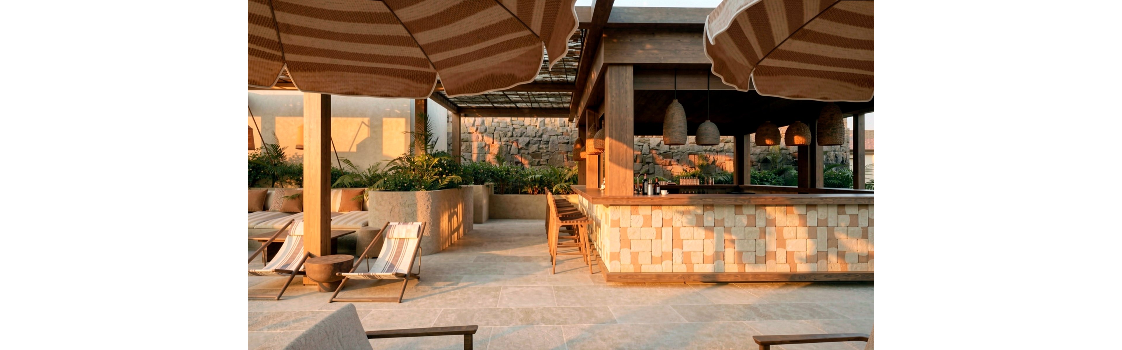 An open-air bar terrace is bathed in warm sunset light, featuring stone and wood construction with a modern rustic design. Lounge chairs and striped sun umbrellas create a relaxed seating area in front of the bar. Hanging woven pendant lights and lush greenery add texture and ambiance to the space. A stone wall backdrop and tiled flooring complete the sophisticated resort setting.