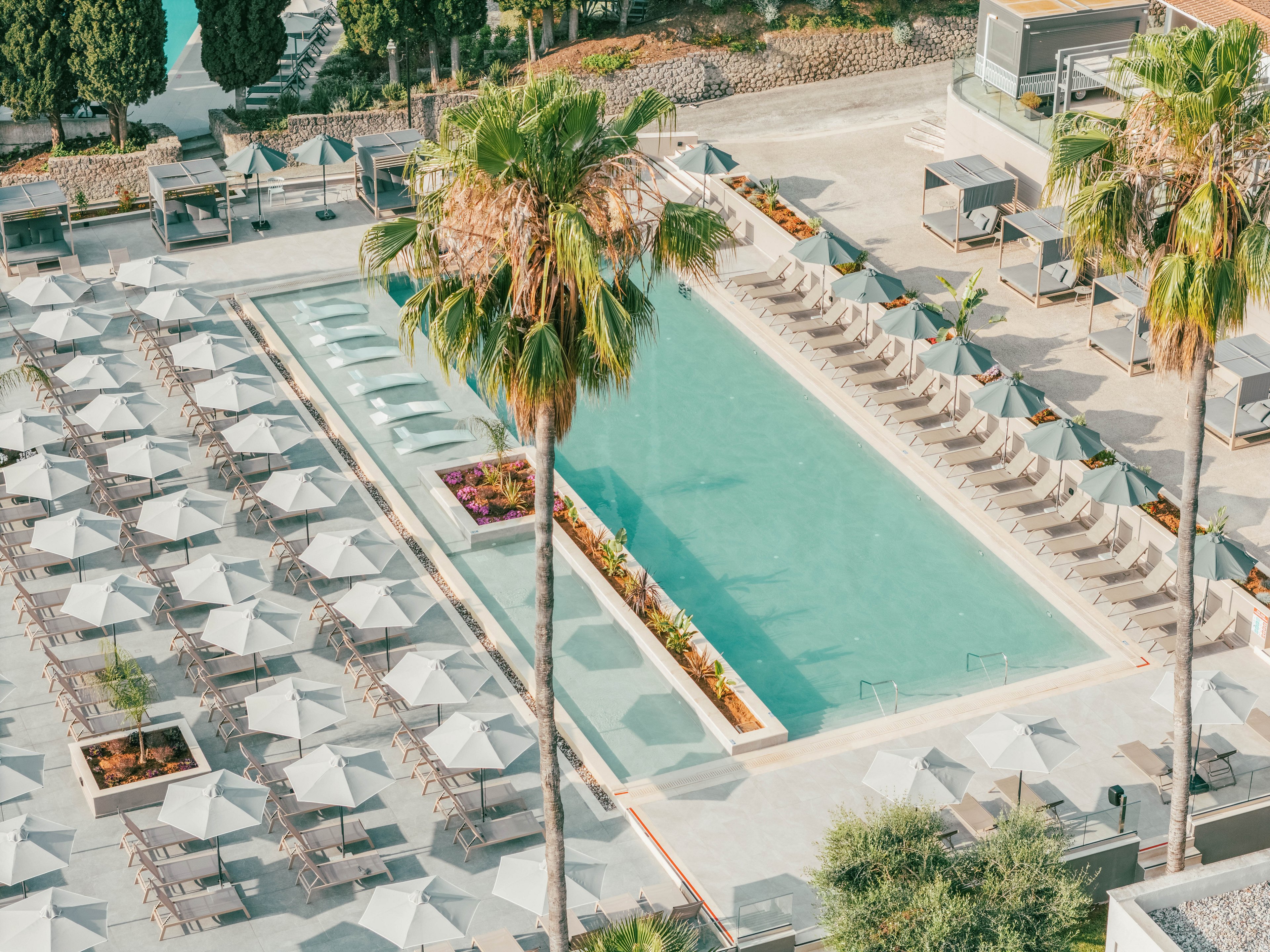 An aerial view showcases a long rectangular swimming pool surrounded by neatly arranged lounge chairs and umbrellas. Tall palm trees frame the pool area, adding a tropical feel to the modern resort setting. The scene appears calm and organized, with cabanas and landscaped planters enhancing the luxurious atmosphere. Soft, muted colors and clear water contribute to a relaxed vacation mood.