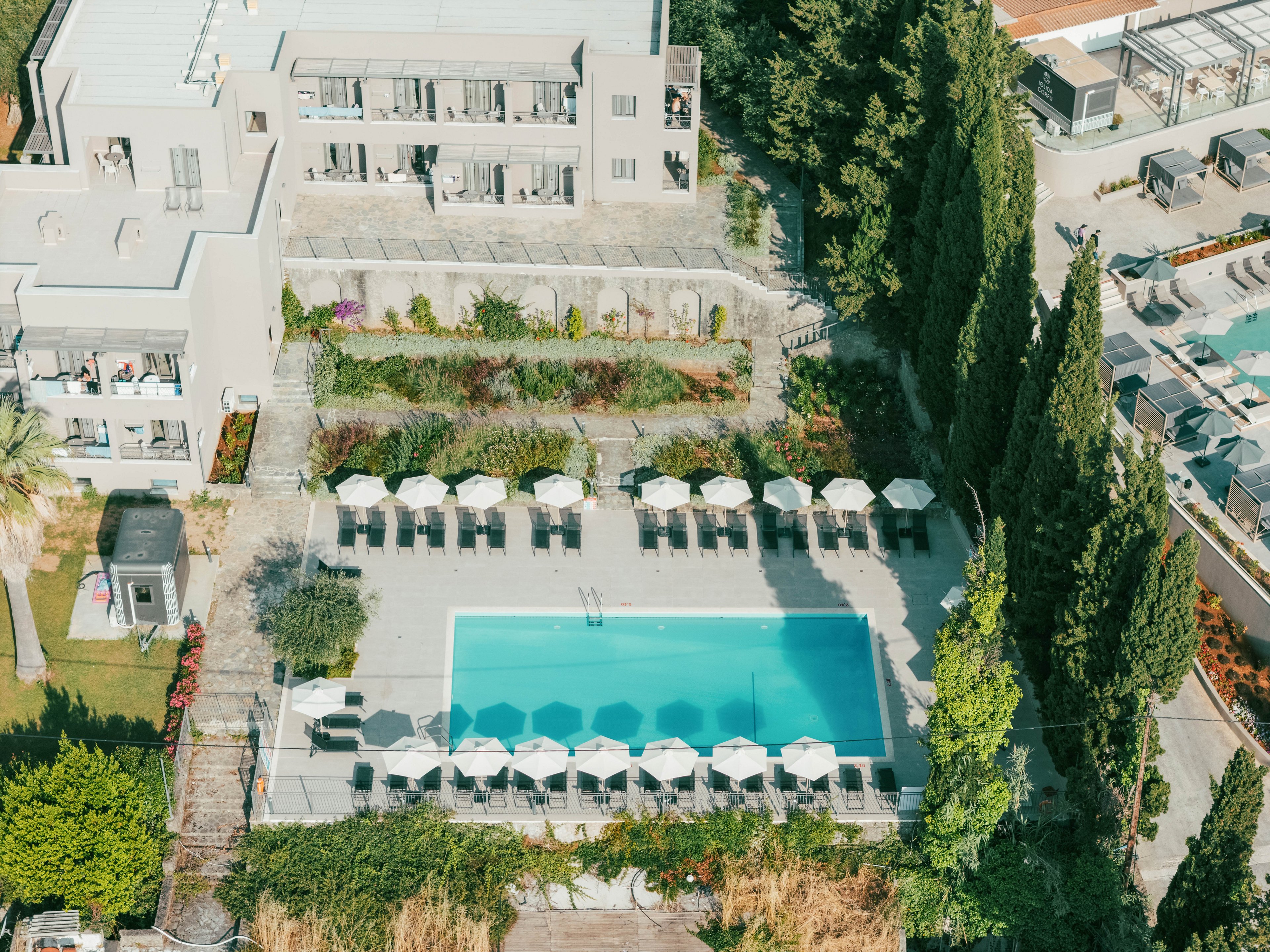An aerial view shows a rectangular outdoor swimming pool at the center of a modern hotel or resort complex. Rows of white sun loungers and umbrellas line both sides of the pool, creating a symmetrical layout. Surrounding the pool are landscaped gardens, tall trees, and contemporary multi-story buildings with balconies. The bright daylight and clear water give the scene a relaxed, vacation atmosphere.