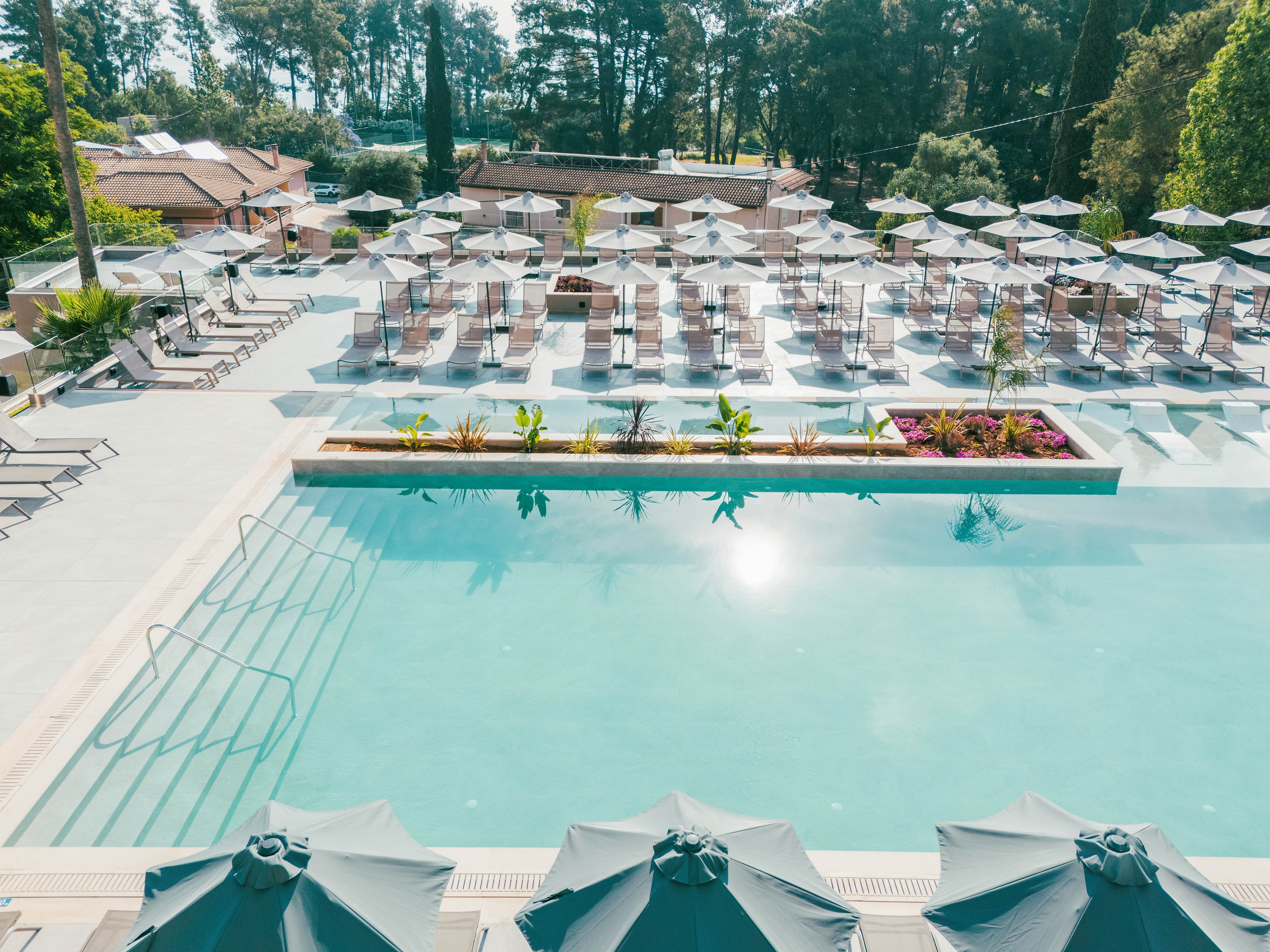 A large outdoor swimming pool is surrounded by neatly arranged sun loungers and umbrellas at a resort. The clear turquoise water reflects the bright daylight, creating a refreshing atmosphere. In the background, tall trees and low buildings frame the pool area, adding a natural backdrop. The scene appears calm and empty, suggesting an early morning or off-peak time at the resort.