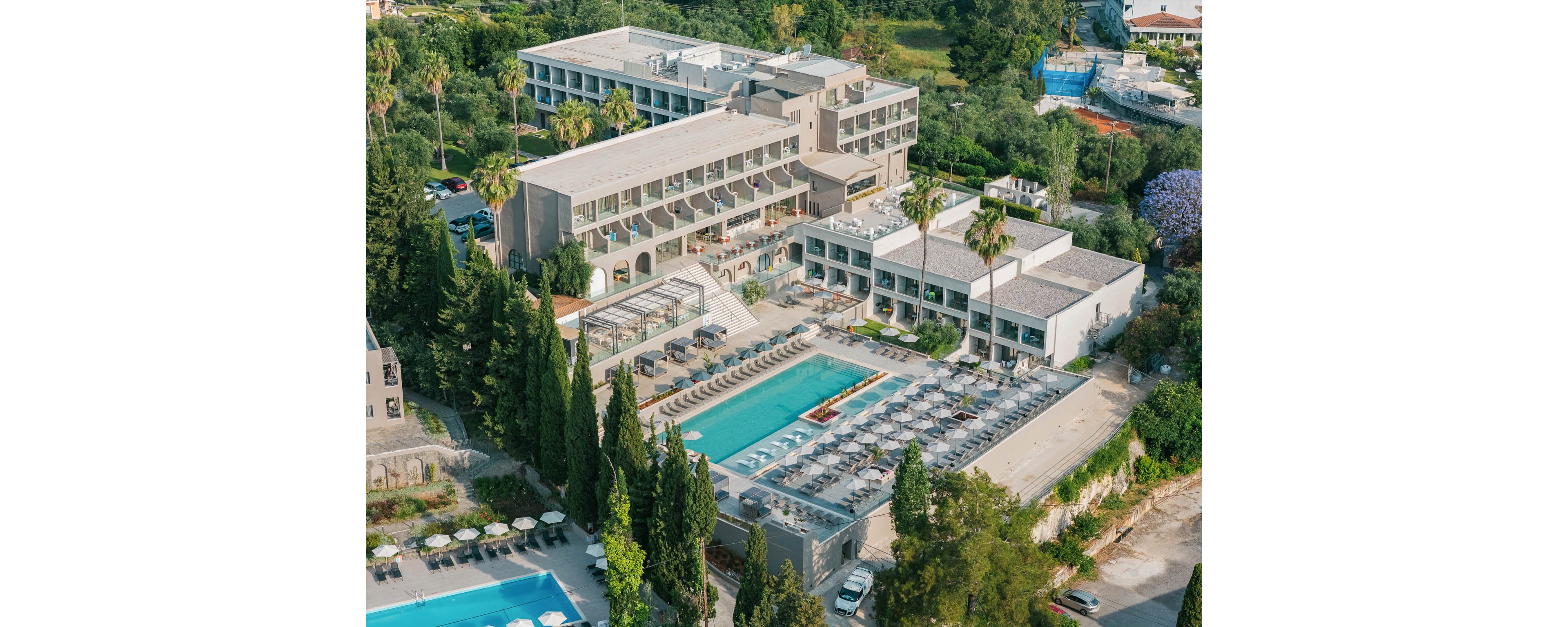 An aerial view showcases a contemporary resort hotel complex surrounded by lush greenery and tall trees. The central focus is a large rectangular swimming pool lined with sun loungers and umbrellas, with additional seating terraces and outdoor areas. Multiple interconnected buildings with flat roofs and balconies frame the pool, suggesting guest rooms and shared amenities. A second pool is visible in the foreground, emphasizing the leisure and vacation atmosphere of the property.