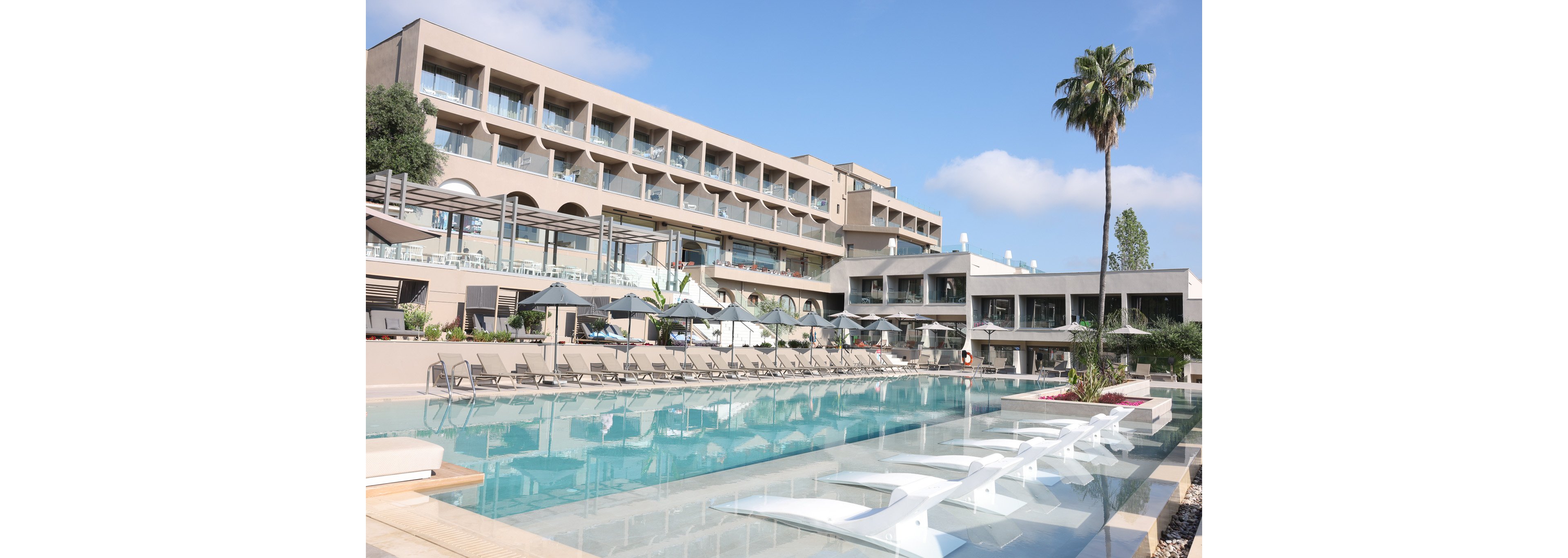 A contemporary multi-story resort hotel overlooks a large outdoor swimming pool. Numerous lounge chairs and umbrellas line the poolside, suggesting a relaxing vacation setting. The architecture features clean lines, glass balconies, and neutral tones under a clear blue sky. A single tall palm tree and landscaped details add a tropical, tranquil atmosphere.
