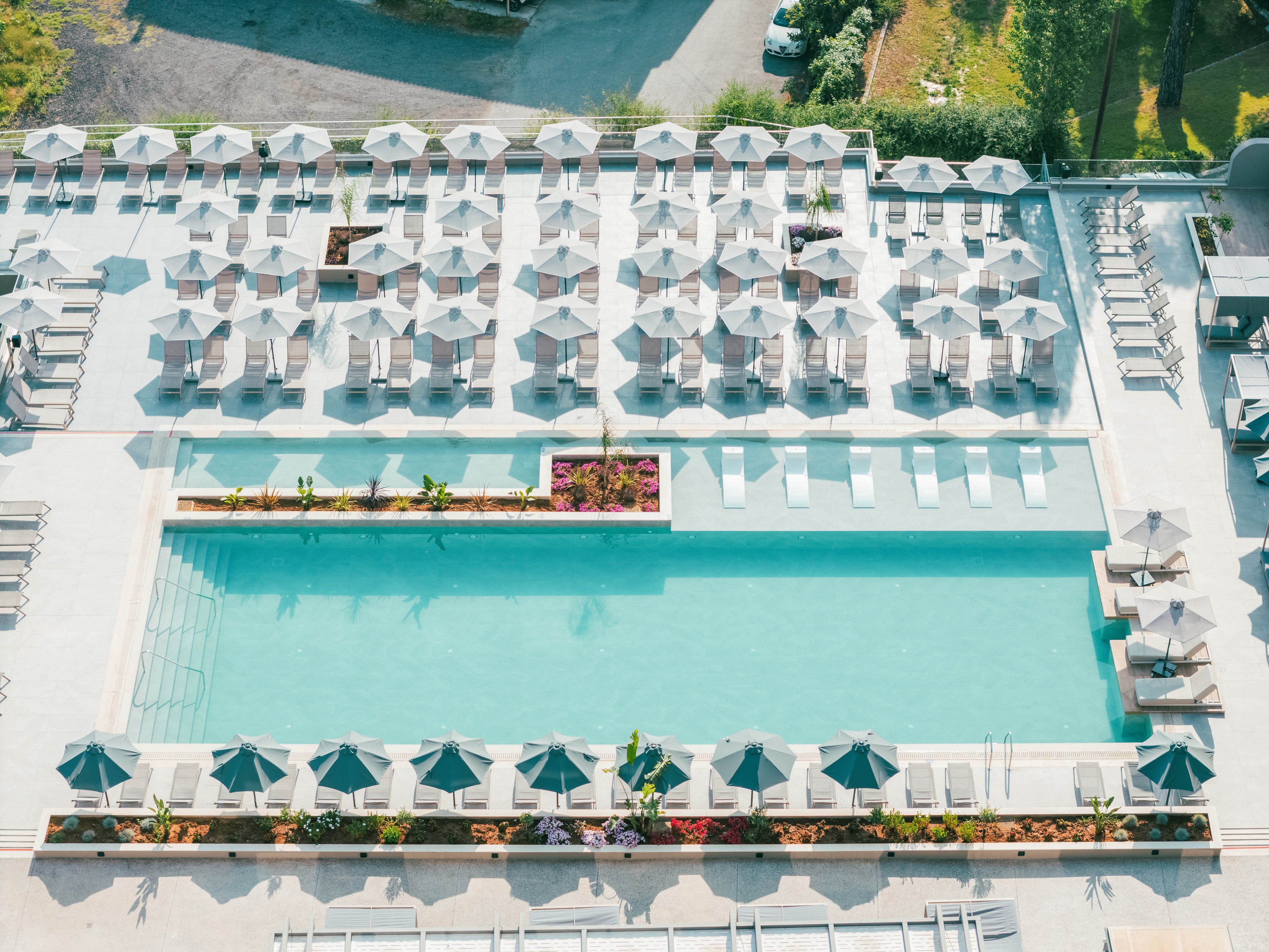 An aerial view shows a large rectangular swimming pool at a modern resort. Numerous sun loungers and neatly arranged umbrellas surround the pool in a symmetrical pattern. A narrow planter with flowers and greenery divides sections of the pool area, adding color to the pale stone deck. The scene appears calm and orderly, suggesting a quiet, sunny day at a vacation destination.