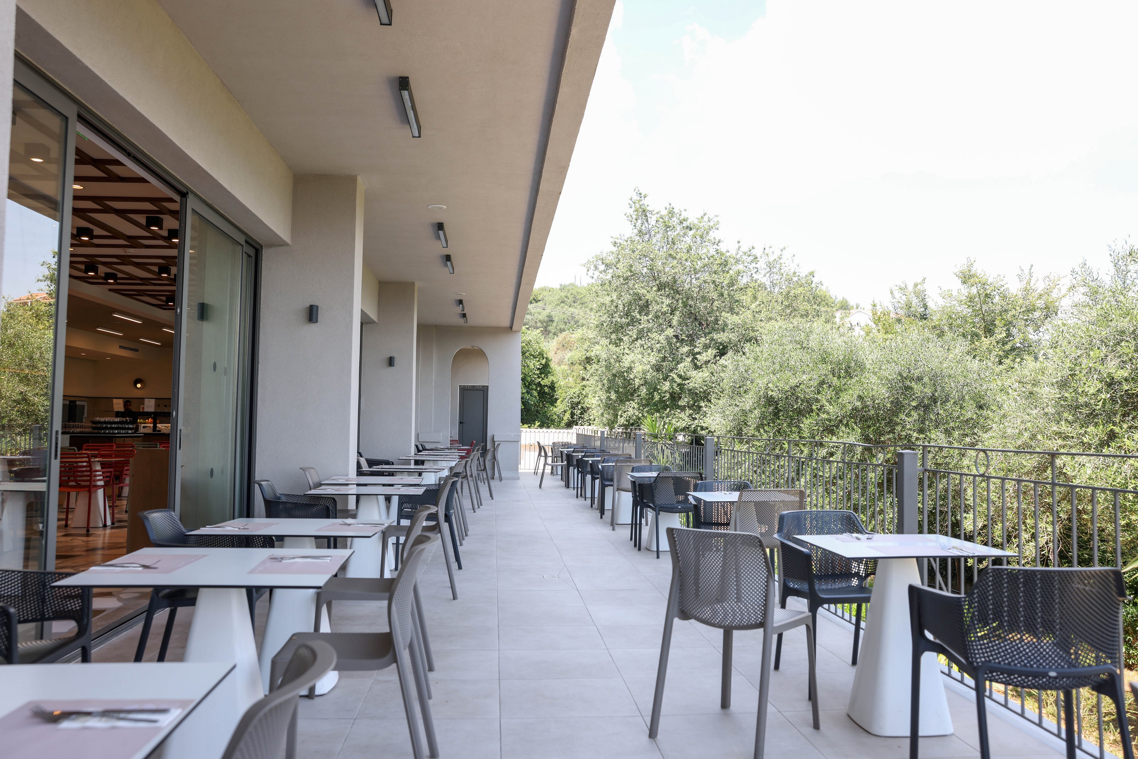 A modern restaurant terrace features neatly arranged tables and chairs along a long balcony. The space is adjacent to an indoor dining area visible through large glass doors. Metal railings line the edge of the terrace, overlooking lush green trees. The scene is bright and quiet, suggesting a relaxed outdoor dining experience.