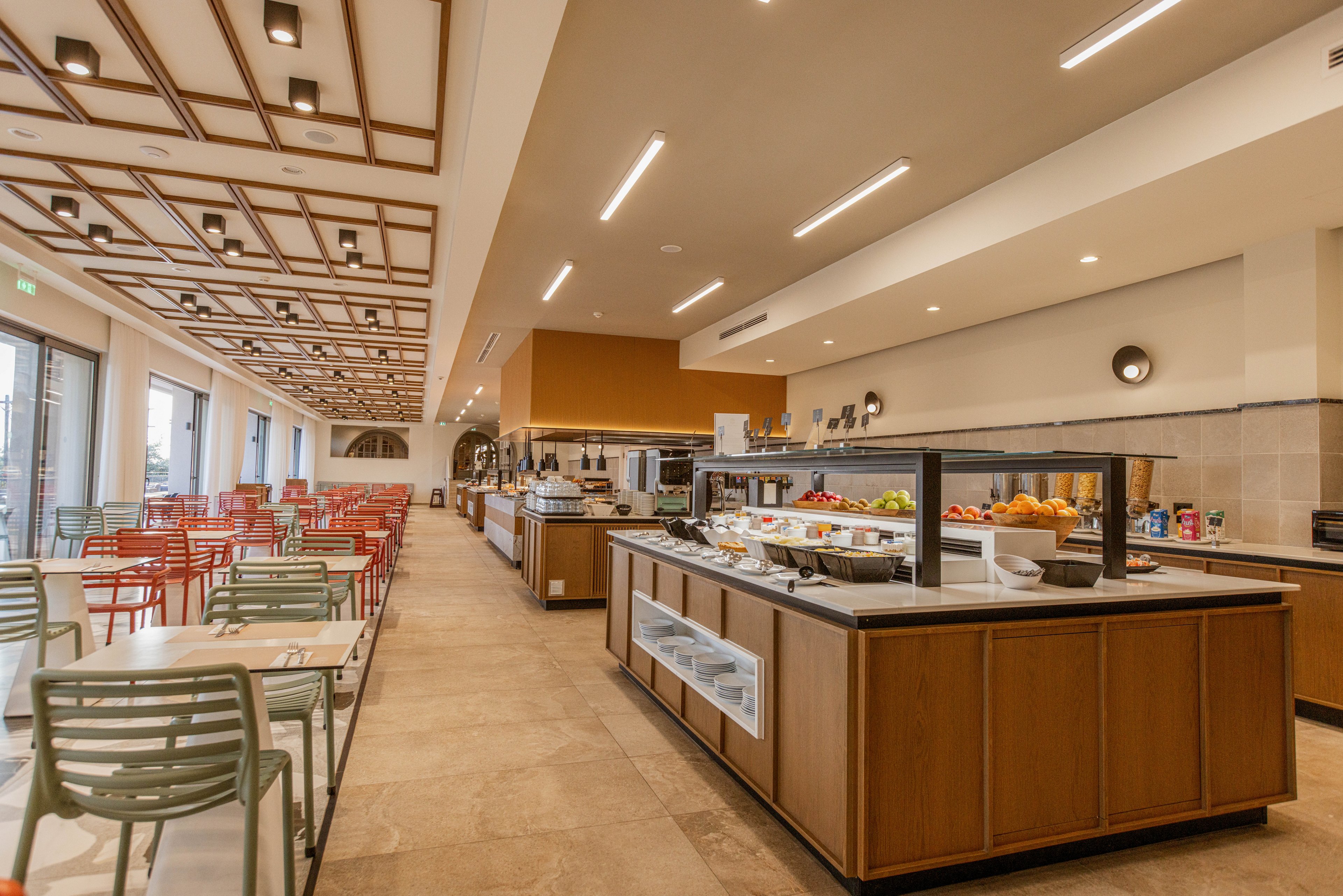 A spacious modern cafeteria features a central self-service buffet counter filled with fruit, salads, and other food items. Rows of colorful chairs and tables line the left side of the room, creating an inviting dining area. Warm lighting, wooden accents, and a clean tiled floor contribute to a contemporary, welcoming atmosphere. Large windows on one side suggest natural light and an open, airy setting.