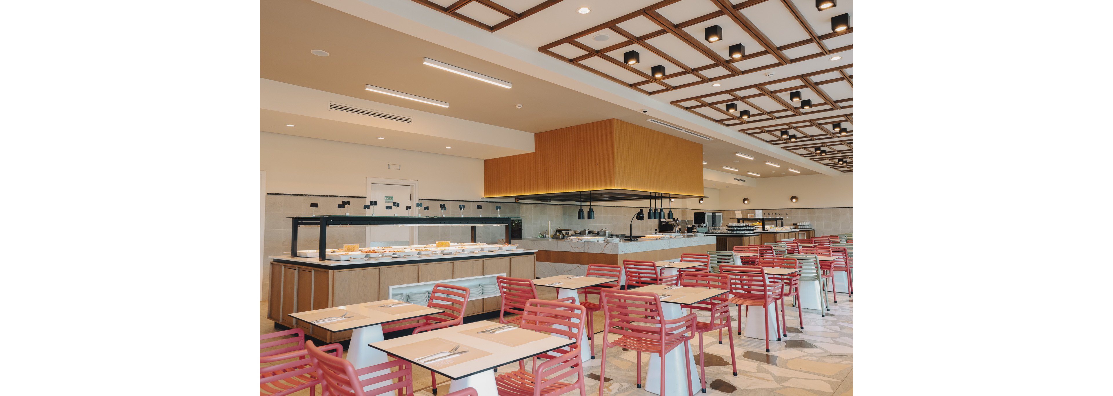A contemporary cafeteria interior features rows of empty tables with red chairs arranged neatly. In the background, a self-service buffet counter displays food trays under warm lighting and heat lamps. The ceiling has a geometric wooden grid design with recessed lighting, creating a bright and orderly atmosphere. The overall setting appears clean, spacious, and ready for diners.