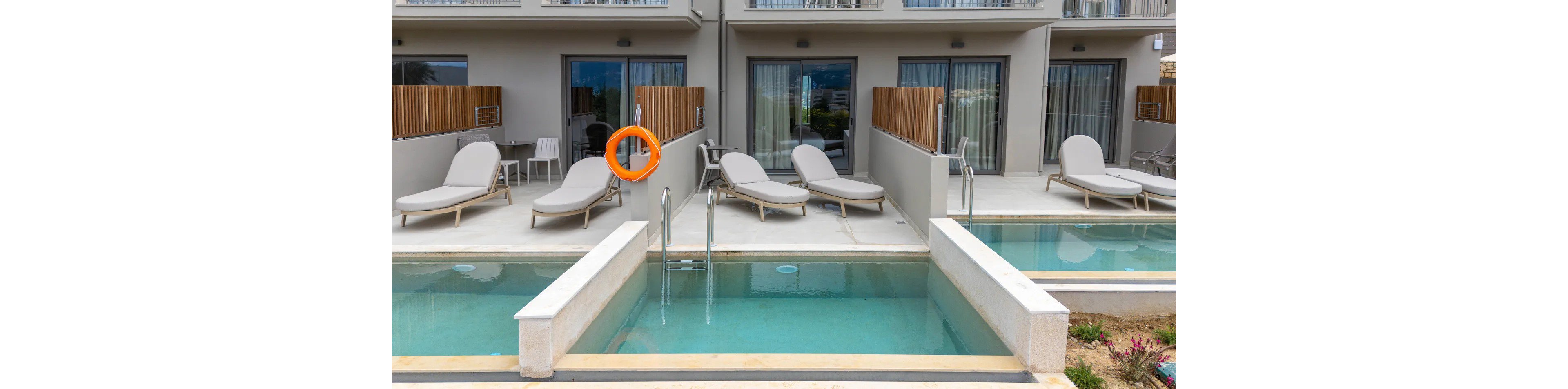 A contemporary hotel terrace features several private plunge pools directly in front of ground-floor rooms. Each patio area includes cushioned sun loungers and small side tables arranged for relaxation. Glass doors and curtains lead into the rooms, while minimalist railings and neutral tones emphasize the modern design. A bright orange life ring hangs on the wall near the pools, adding a safety element and a pop of color.