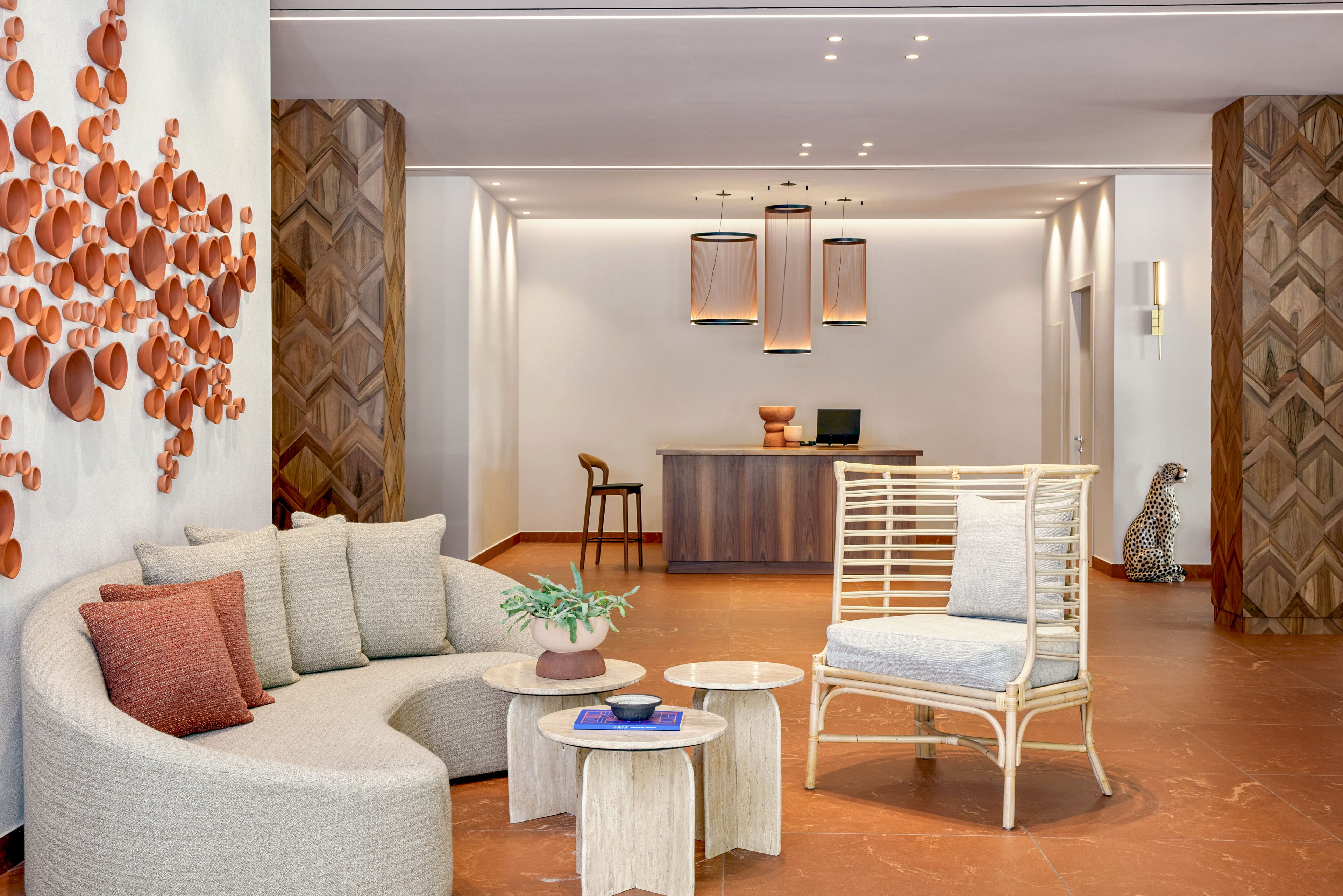 A stylish hotel lobby features a curved sofa and a woven armchair arranged around small wooden coffee tables. The space is bright and minimal, with warm terracotta flooring and wood-paneled columns framing a simple reception desk in the background. Decorative wall art made of circular elements adds texture to the left wall, while pendant lights hang above the desk area. A sculptural animal figure stands near the right wall, contributing to the eclectic, modern interior design.