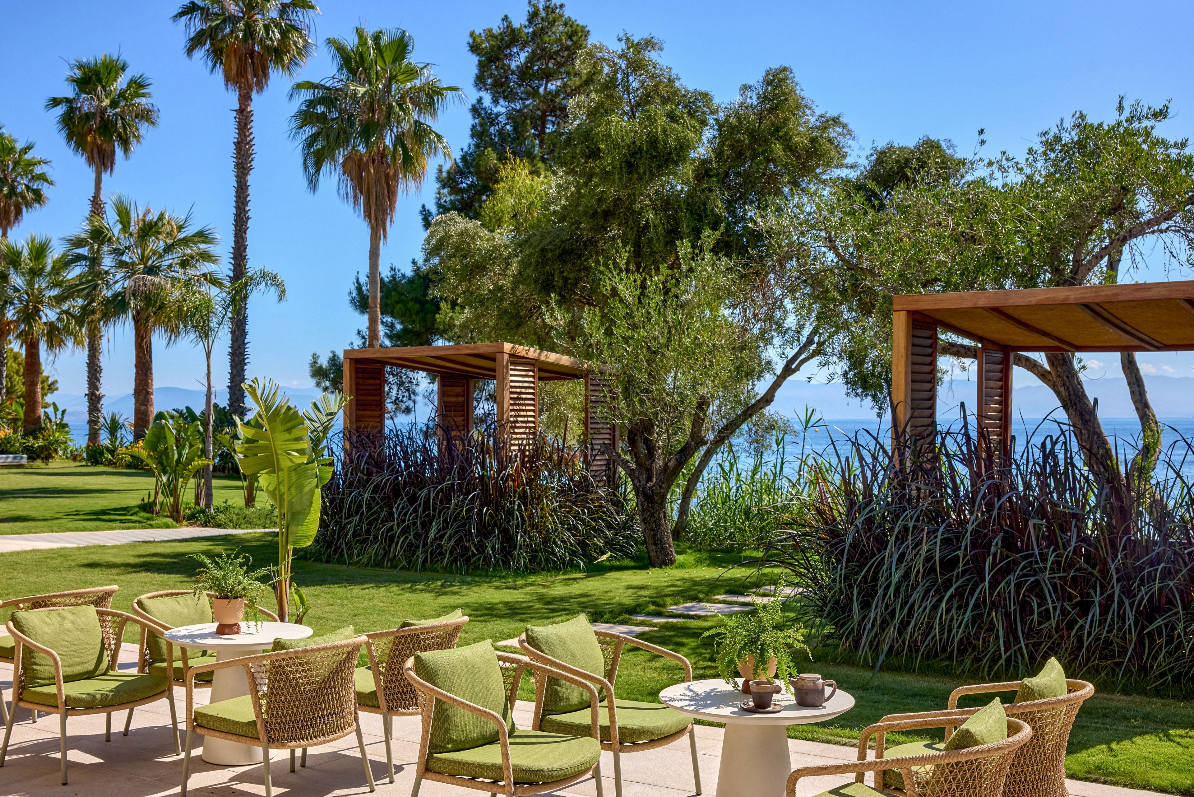 An inviting outdoor lounge area features green-cushioned chairs and small tables arranged on a sunny patio. Wooden cabanas and lush landscaping with tall palm trees and dense shrubs frame the space. In the background, the blue sea and clear sky create a tranquil coastal setting. The overall scene suggests a relaxing resort environment ideal for leisure and socializing.
