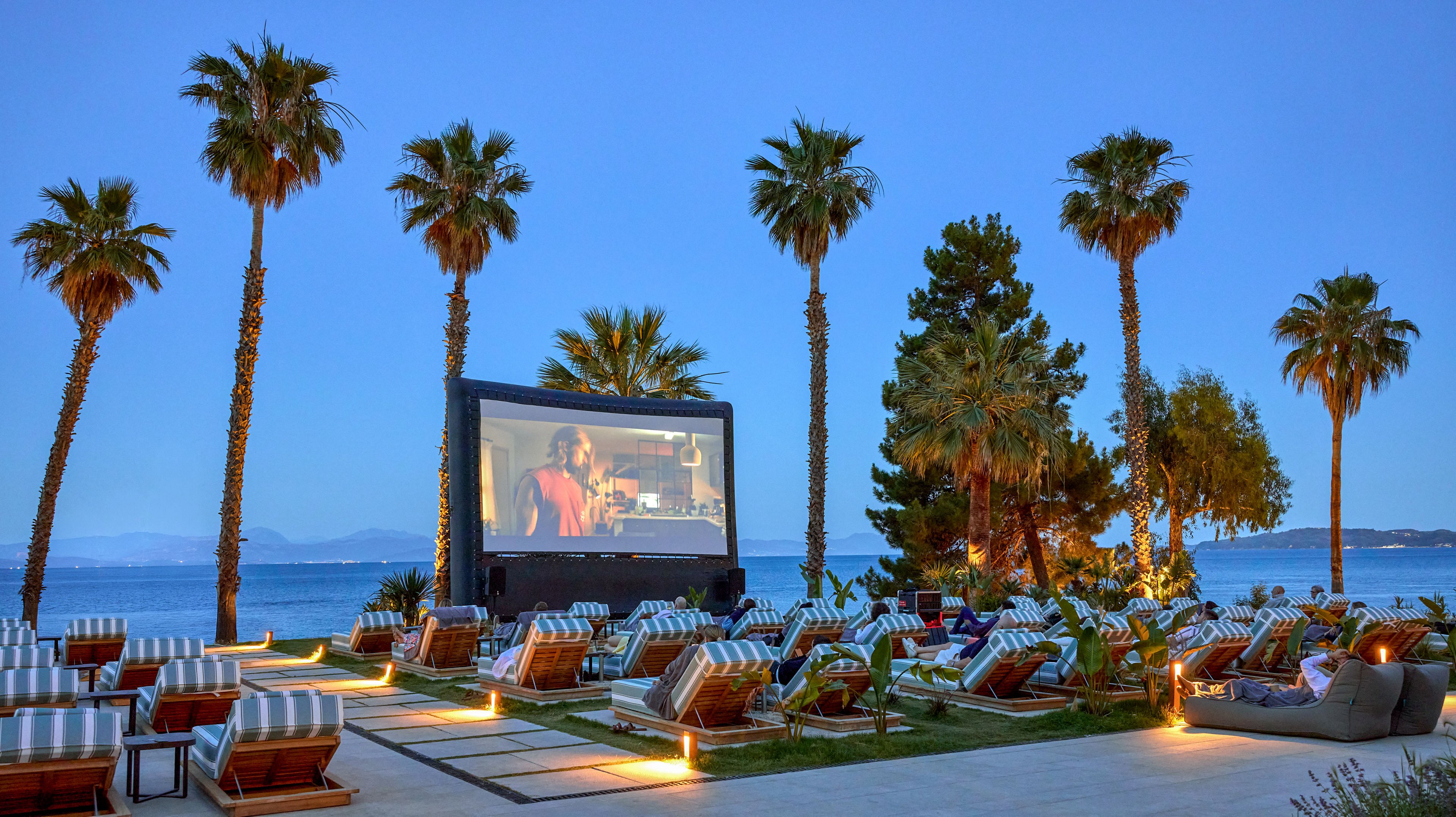 An outdoor cinema screen is set up on a lawn overlooking a calm sea at dusk. Rows of cushioned lounge chairs and striped sunbeds face the large screen, creating a relaxed viewing area. Tall palm trees frame the scene, and subtle ground lighting adds warmth to the twilight atmosphere. A few people are seated, enjoying the movie in this resort-like beachfront setting.