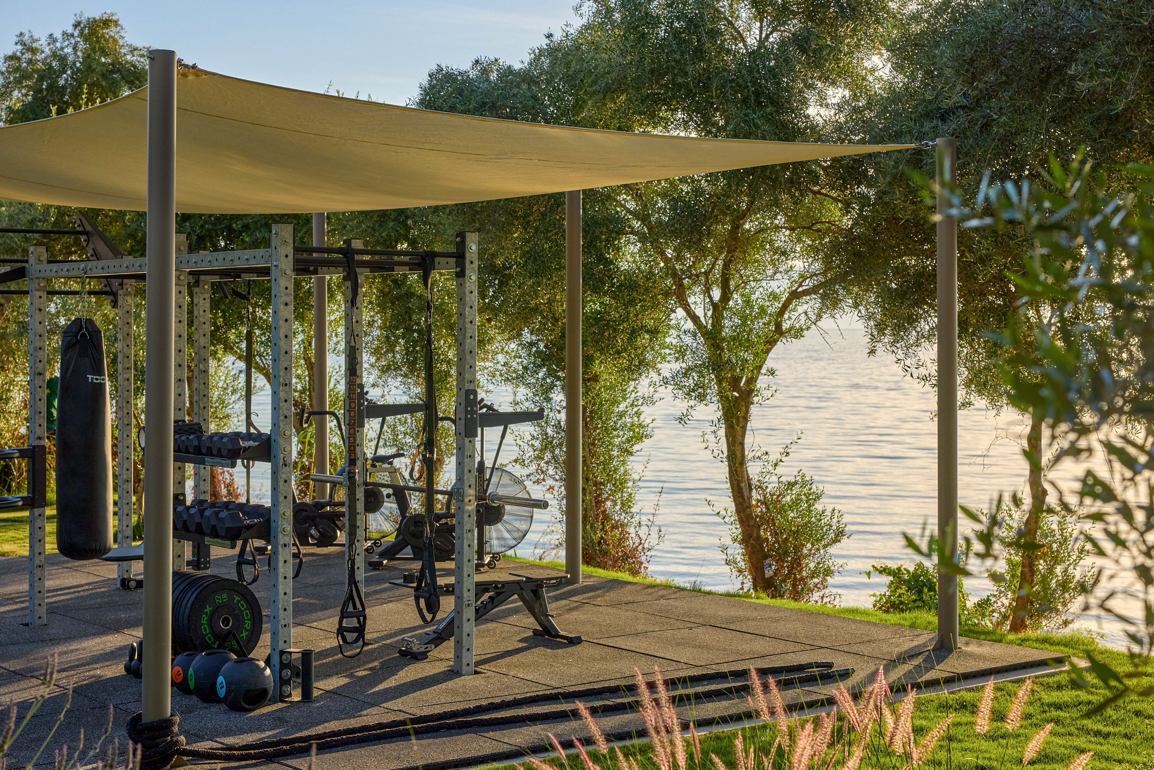 An outdoor gym setup stands on a shaded platform near a calm waterfront. The structure includes weight racks, barbells, plates, and benches arranged neatly under a fabric canopy. Lush green grass, trees, and plants surround the exercise area, creating a natural and tranquil environment. Soft sunlight reflects off the water, giving the fitness space a peaceful, scenic atmosphere.