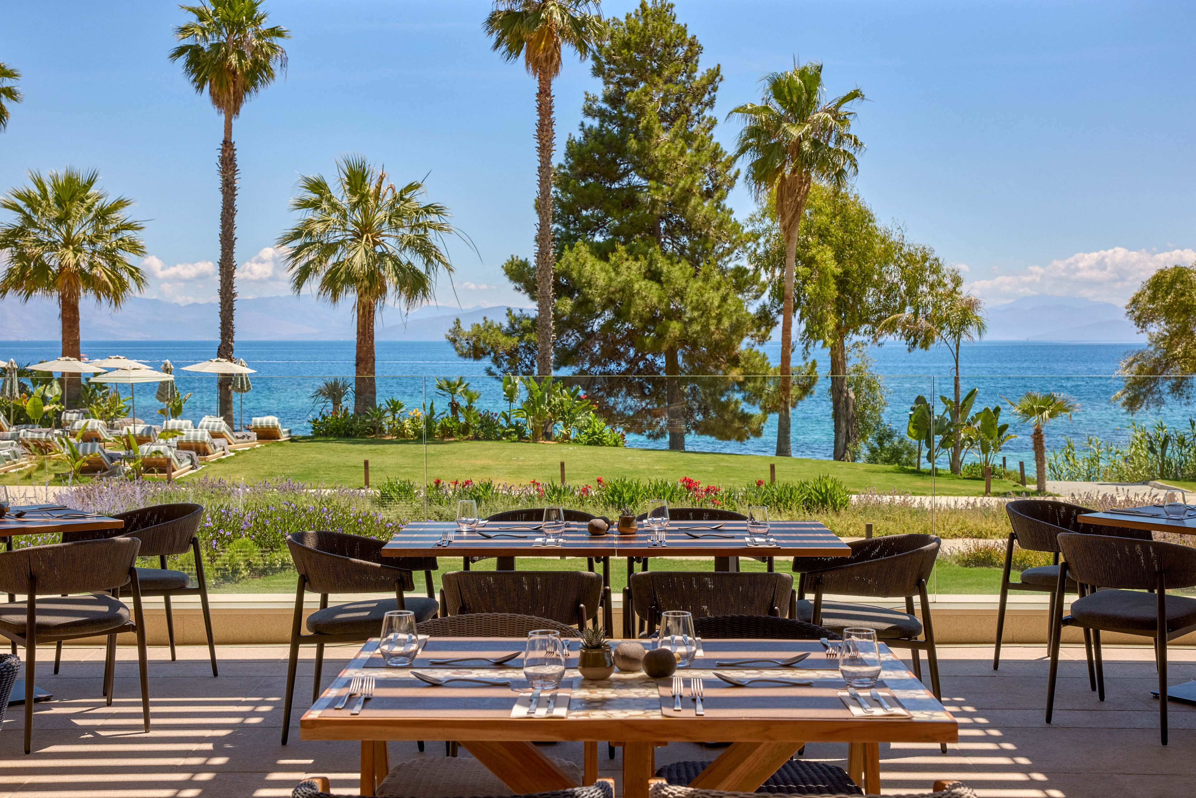 An elegant outdoor restaurant terrace overlooks a lush garden and a calm blue sea. Wooden tables are neatly set with glassware and cutlery, ready for guests. Palm trees and other greenery frame the view, leading the eye toward the distant coastline and mountains. Sunlit seating and clear skies create a relaxed, vacation-like atmosphere.