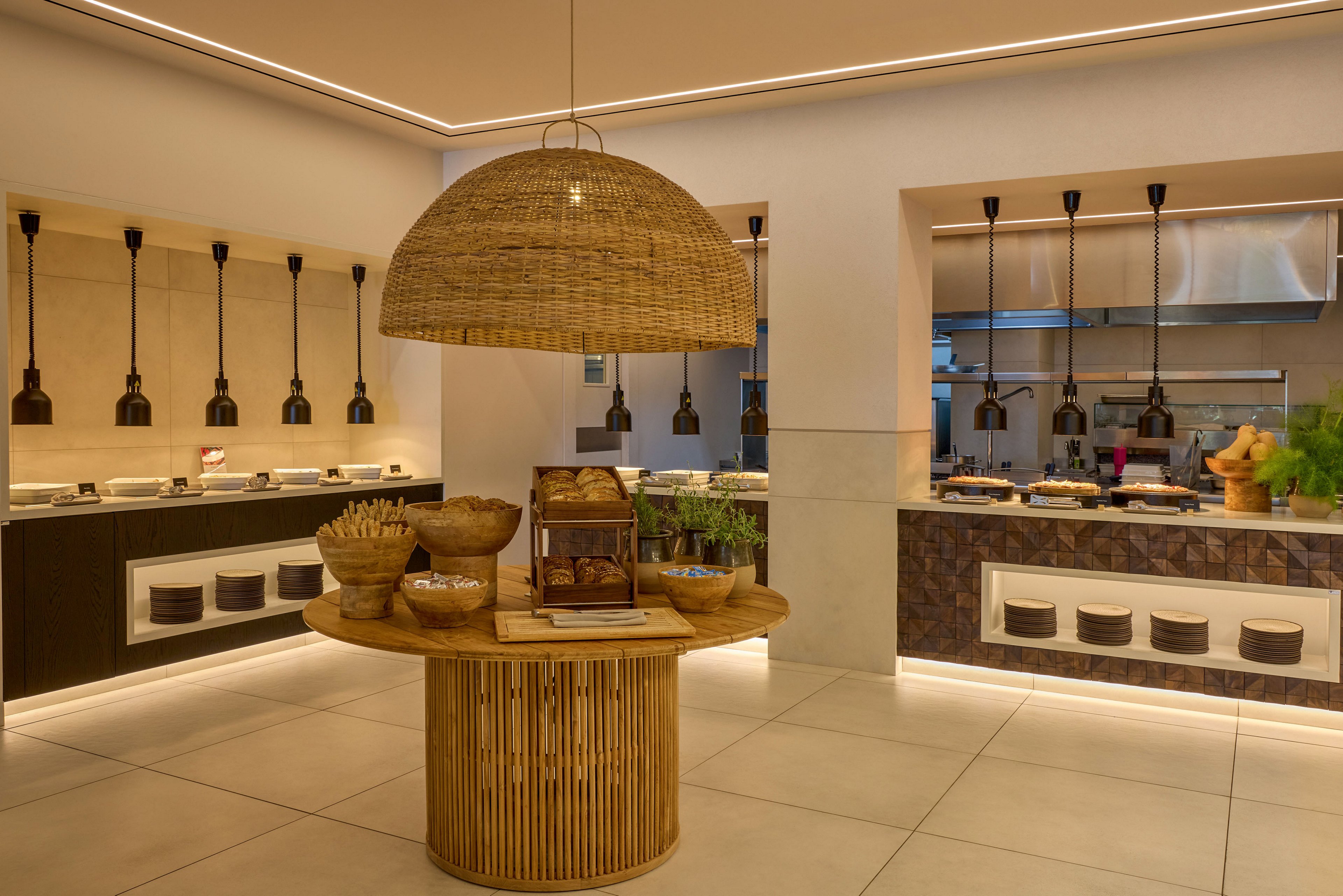 A contemporary buffet restaurant interior features a central round wooden island table with baskets and food displays under a large woven pendant lamp. In the background, long counters present various dishes in metal chafing trays, accented by hanging industrial-style lights. The space is finished with light tiled flooring, clean white walls, and subtle recessed ceiling lighting that creates a warm, inviting atmosphere. Natural materials and neutral tones give the setting a modern yet cozy dining experience.