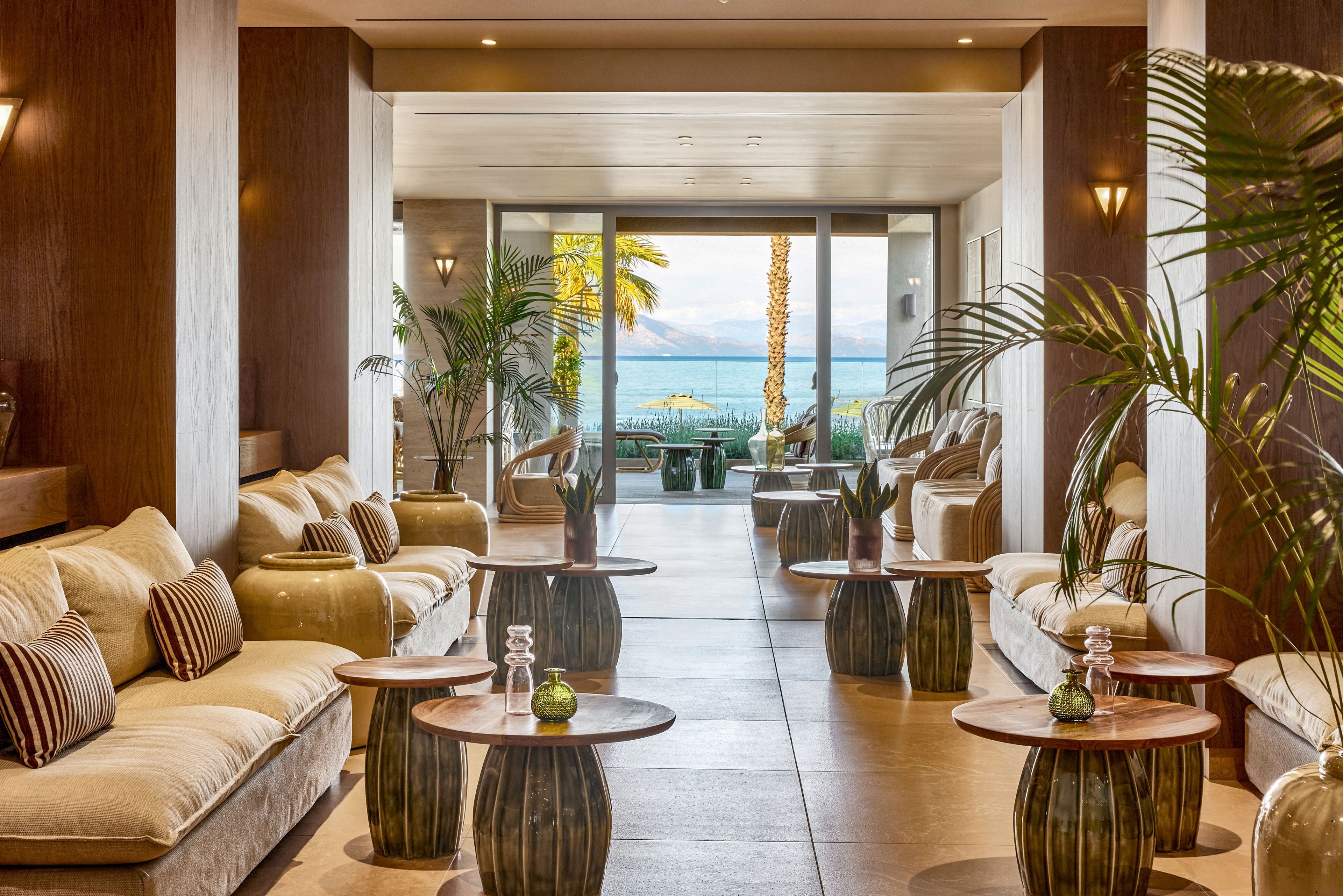 Upscale lounge area with plush beige sofas and striped cushions opens toward a terrace overlooking the sea. Wooden side tables with small green plants line the central walkway, creating a symmetrical and inviting space. Tall palm trees and outdoor seating are visible through large glass doors, framing the coastal scenery. Warm lighting and natural materials contribute to a relaxed, resort-style atmosphere.