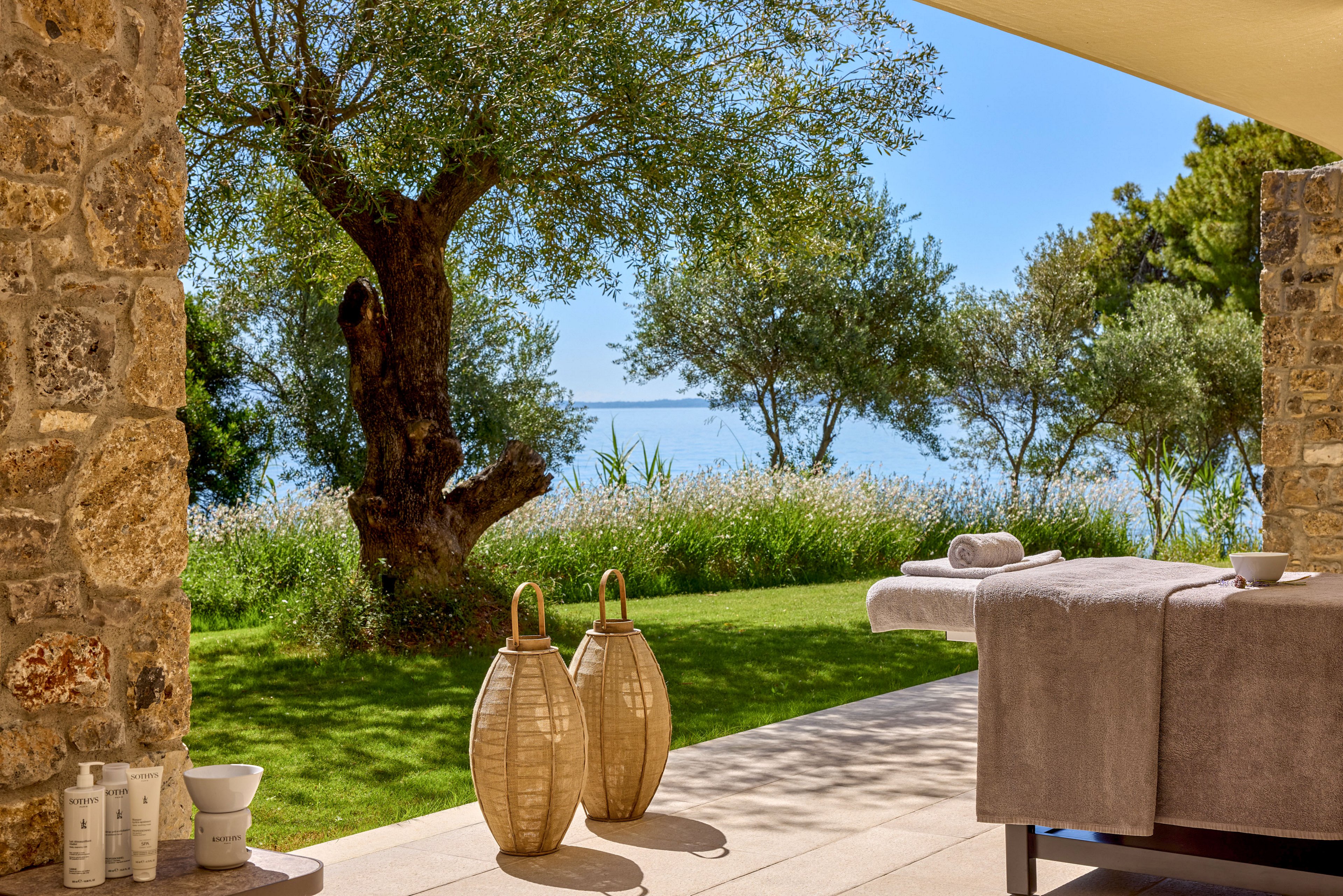 A serene spa terrace opens onto a lush garden with olive trees and a distant view of the sea. A massage table covered in soft gray linens sits under a shaded patio beside stone walls. Decorative lanterns stand on the tiled floor, adding a warm, rustic touch to the scene. Skincare products and bowls are arranged neatly in the foreground, enhancing the relaxing, wellness-focused atmosphere.