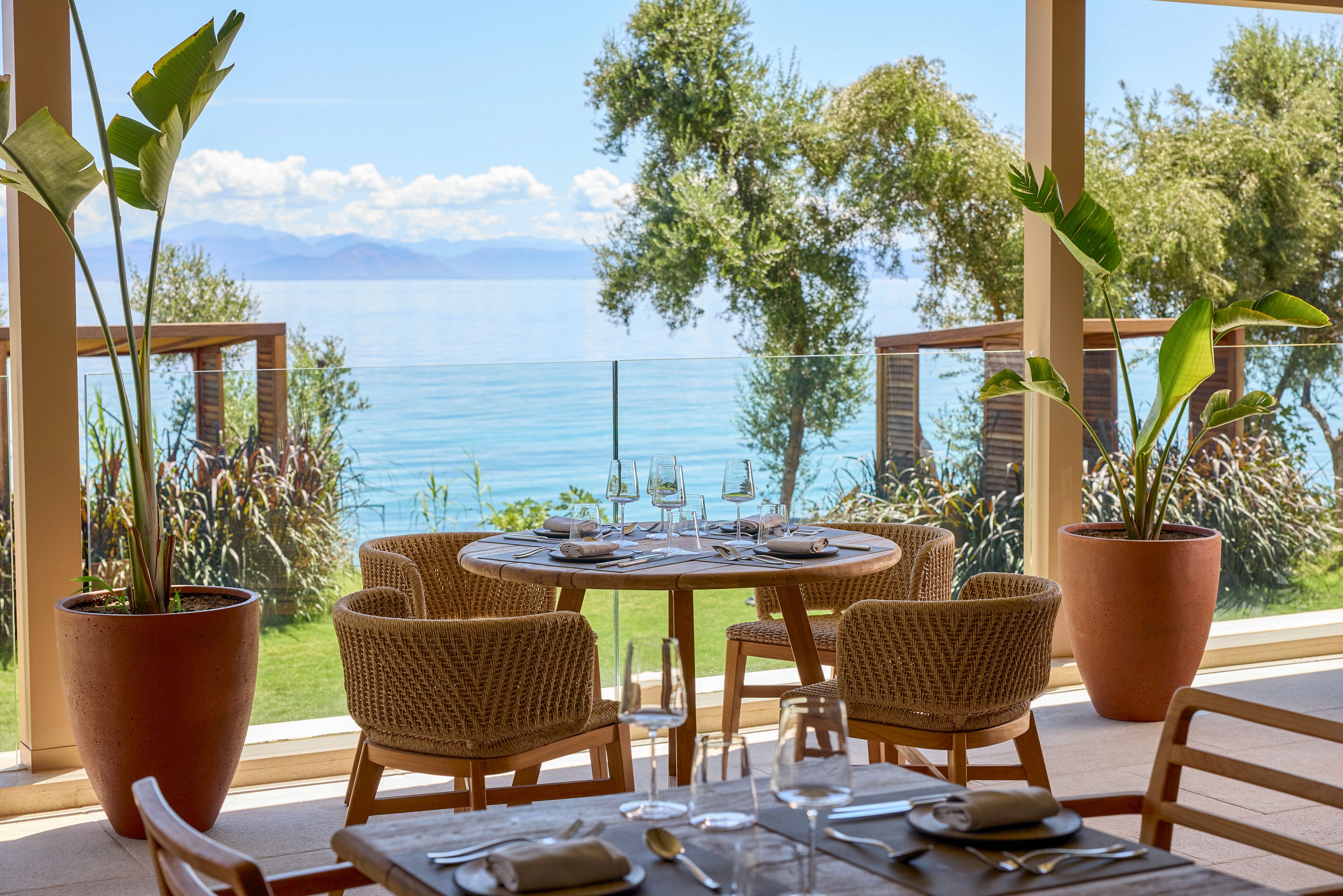 A round wooden dining table is set with glassware and plates on a bright terrace overlooking the sea. Woven chairs surround the table, and large potted plants frame the open view through floor-to-ceiling windows. Lush greenery and trees lead down to the water, with distant mountains under a partly cloudy sky. The scene suggests a relaxed, upscale coastal dining experience in natural light.