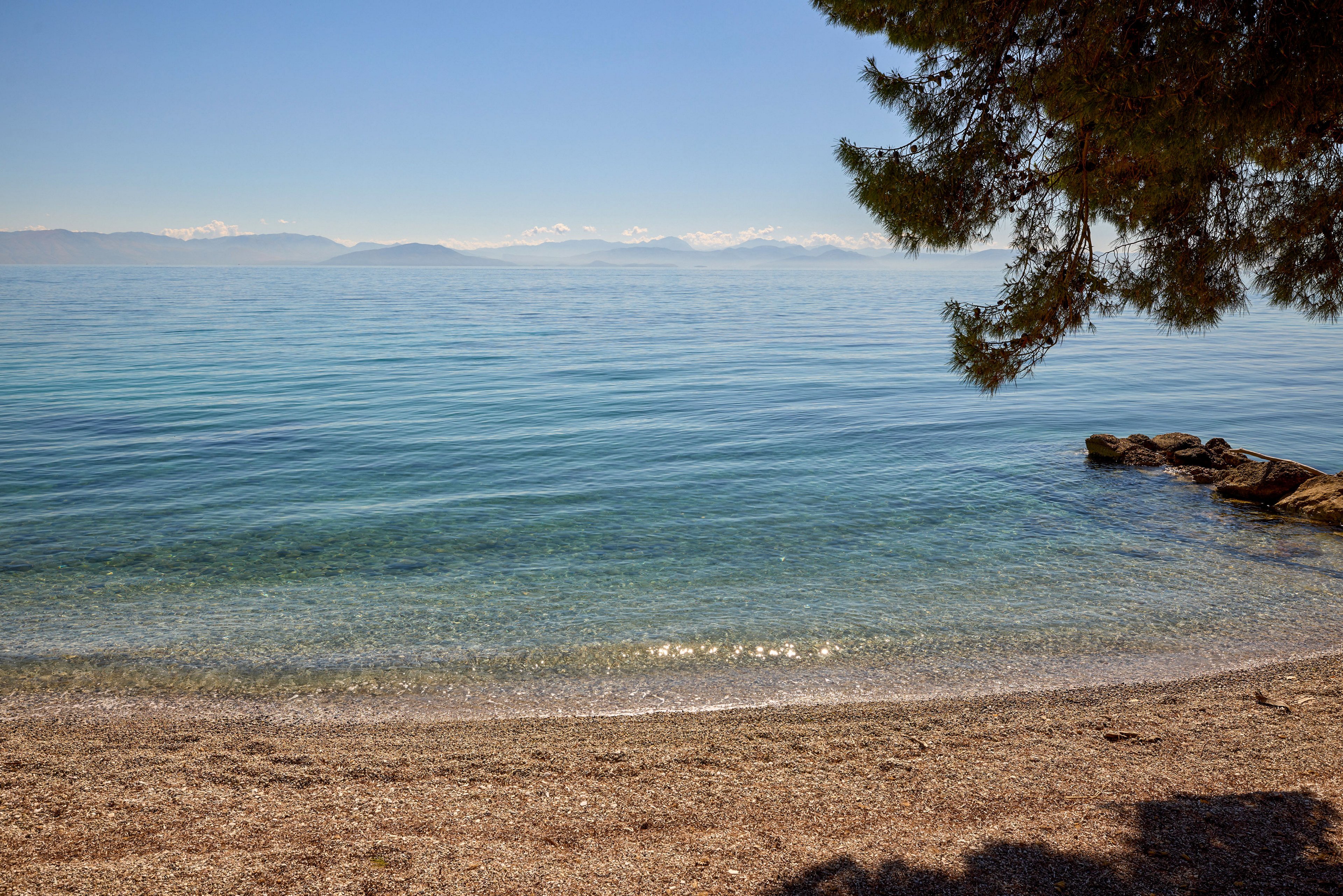 A tranquil pebble beach stretches along the shoreline beside a calm, clear blue sea. Gentle waves lap against the shore under a bright, cloudless sky. Distant mountains are faintly visible across the water, adding depth to the coastal landscape. A tree frames the scene from the right, casting soft shadows on the beach and enhancing the peaceful atmosphere.