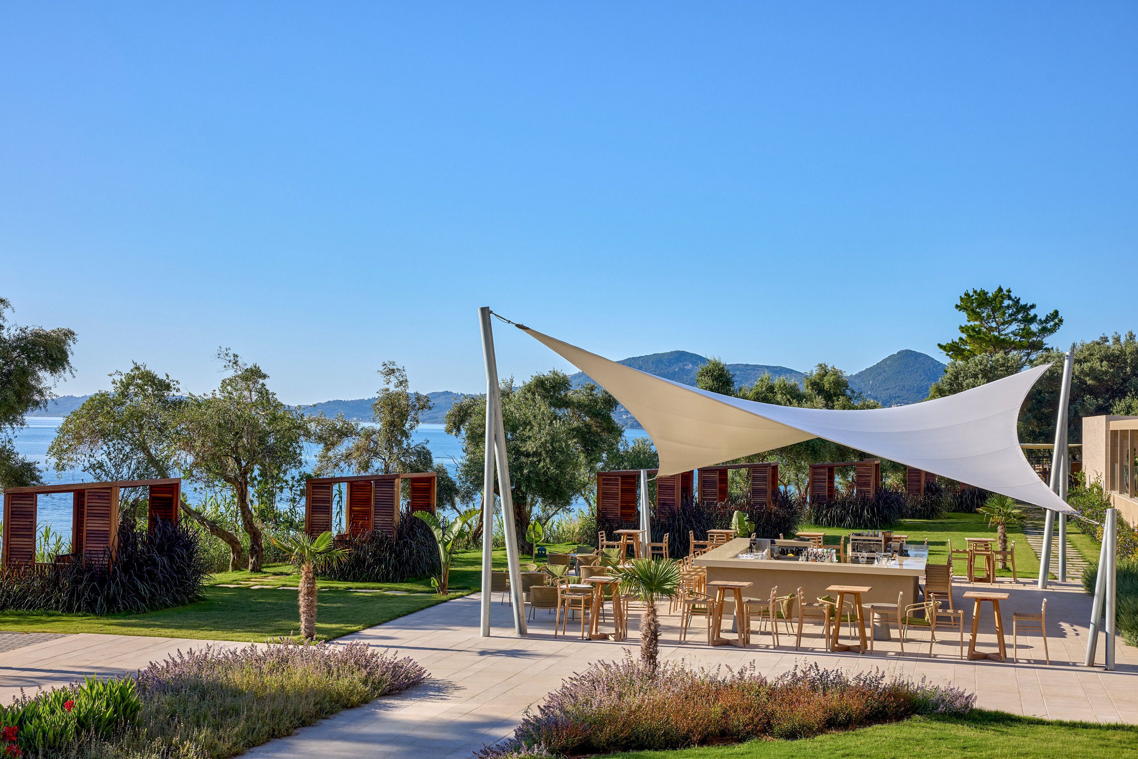 A contemporary open-air bar with wooden stools sits beneath a sleek white shade sail. The terrace is surrounded by manicured lawns, ornamental grasses, and trees, creating a relaxed resort atmosphere. In the background, calm blue water and distant hills frame the coastal setting under a clear sky. The scene suggests leisure, vacation, and social gatherings in a stylish seaside environment.