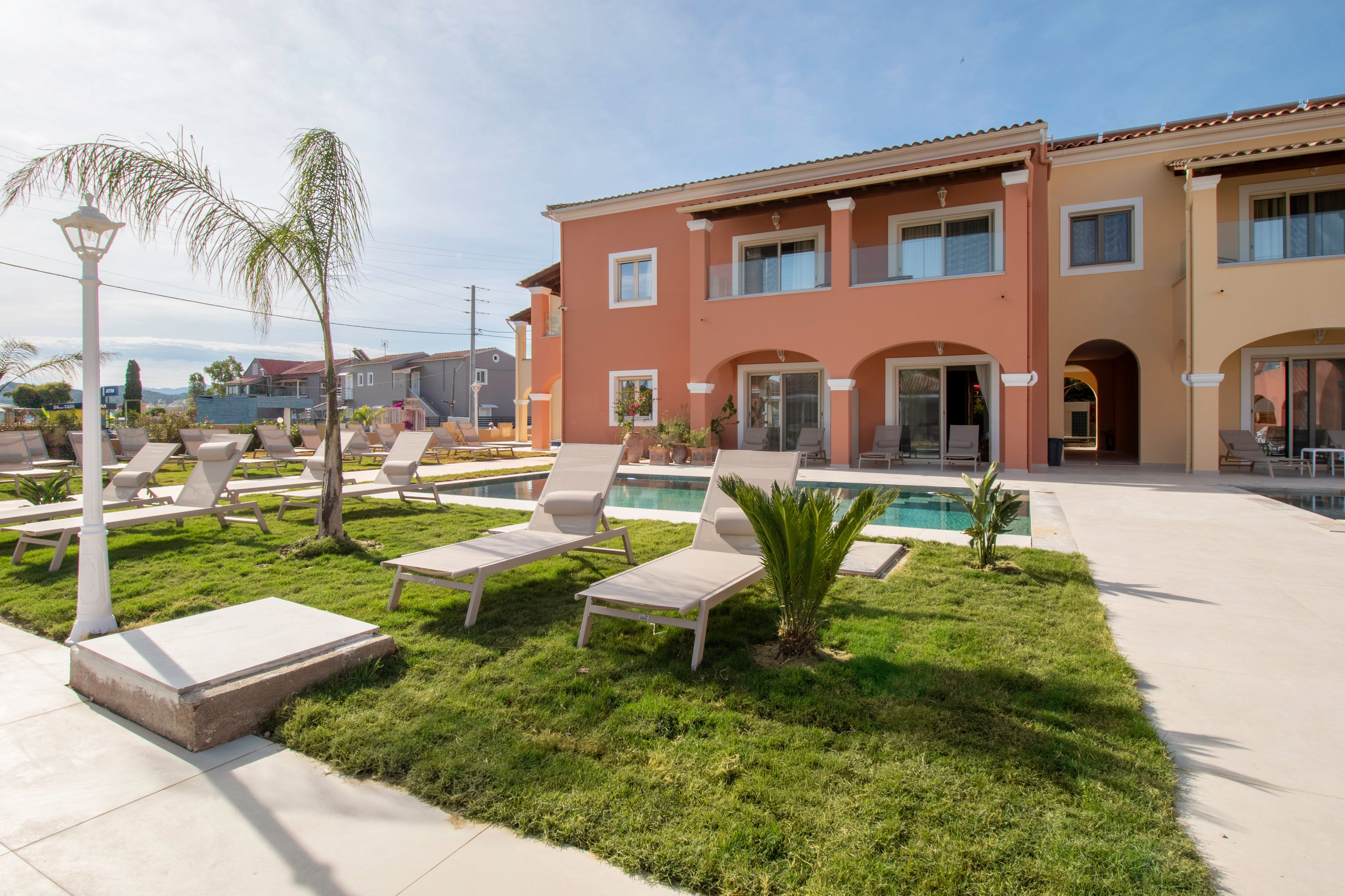 A modern two-story building with warm orange and beige walls overlooks a neatly landscaped courtyard. Several sun loungers are arranged around a small swimming pool on a bright, sunny day. Young palm trees and trimmed grass add a tropical feel to the outdoor space. The scene suggests a quiet resort or hotel area designed for relaxation and leisure.