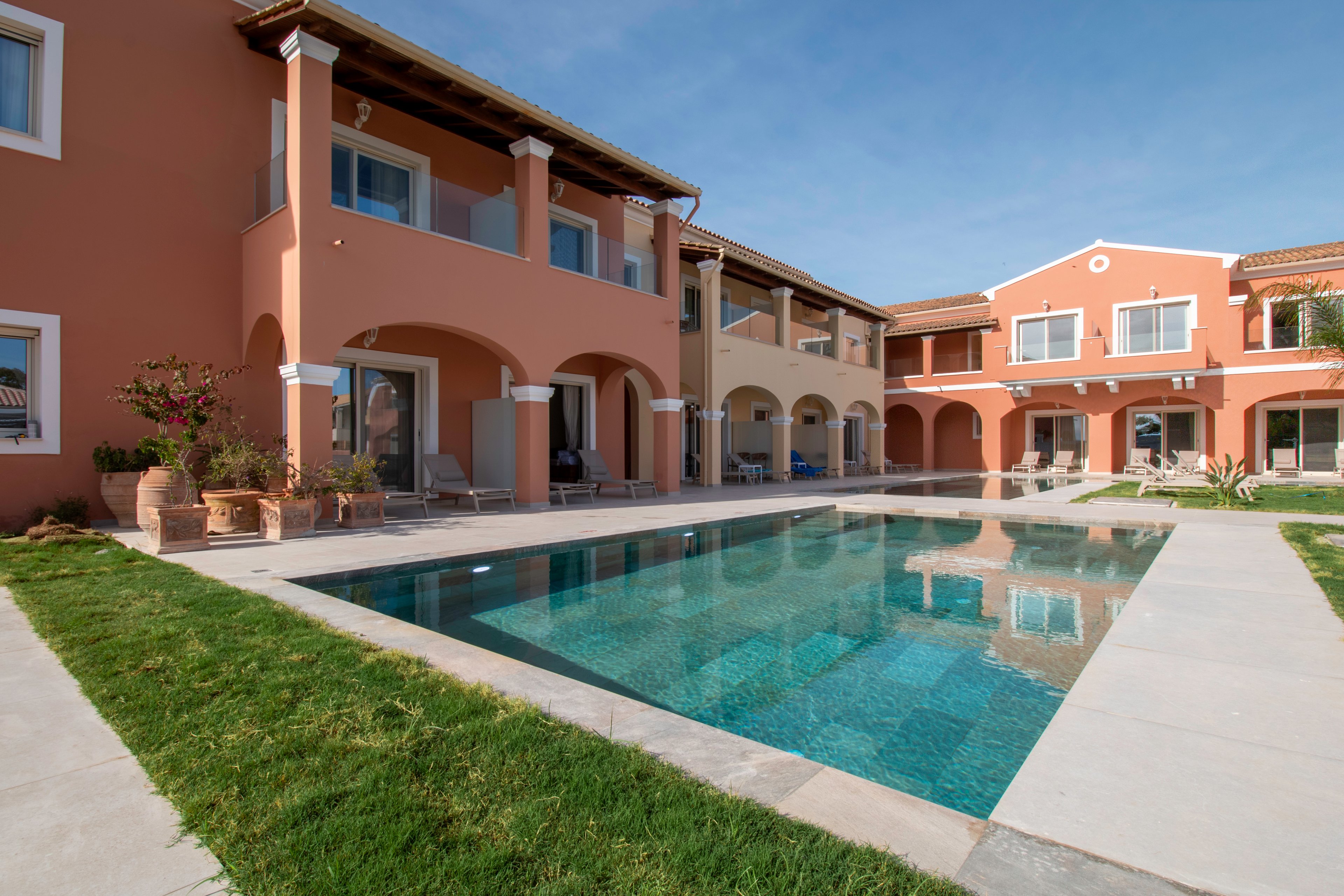 A Mediterranean-style villa complex features a long rectangular swimming pool in the central courtyard. The peach and terracotta colored buildings have arched patios, balconies, and white-framed windows facing the water. Lounge chairs and potted plants line the poolside, suggesting a relaxing resort atmosphere. Clear blue sky and neatly trimmed grass enhance the bright, sunny setting.