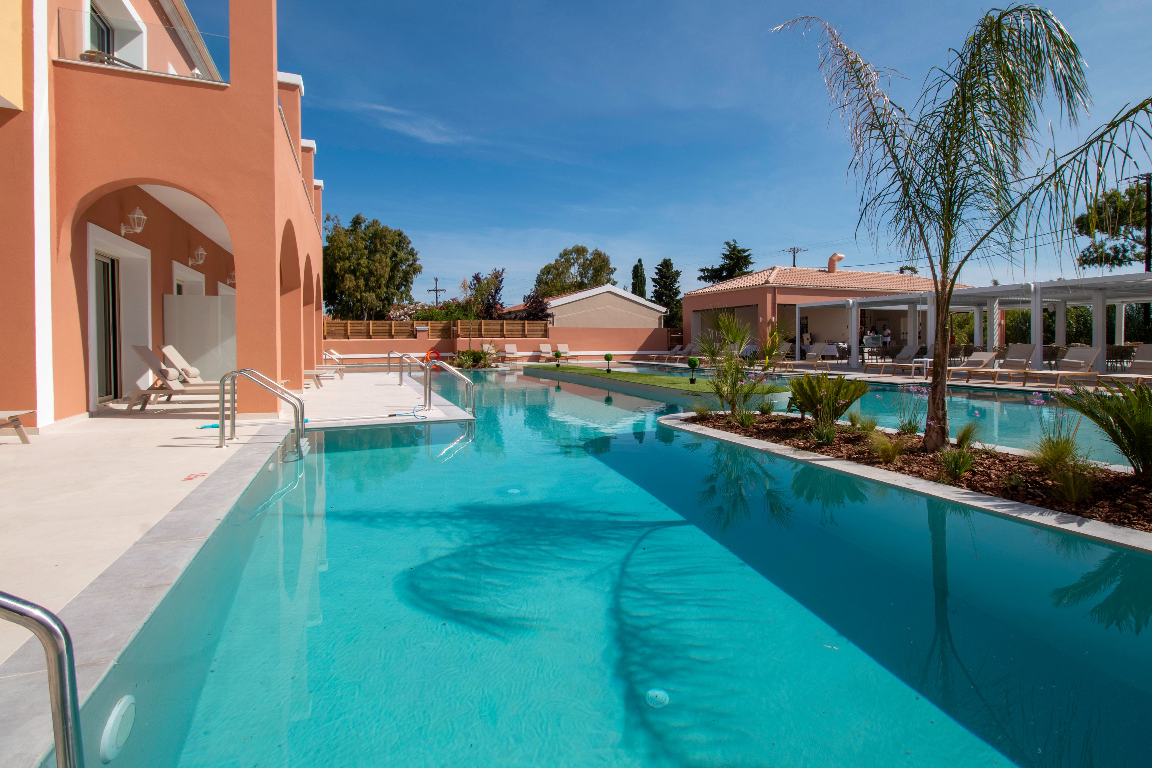 A large, modern swimming pool curves around a peach-colored building under a clear blue sky. Sun loungers and umbrellas line the poolside, suggesting a relaxing resort environment. Palm trees and landscaped greenery decorate the pool’s edge, reflecting in the calm turquoise water. Additional low-rise buildings and covered outdoor areas are visible in the background, enhancing the sense of a spacious vacation property.