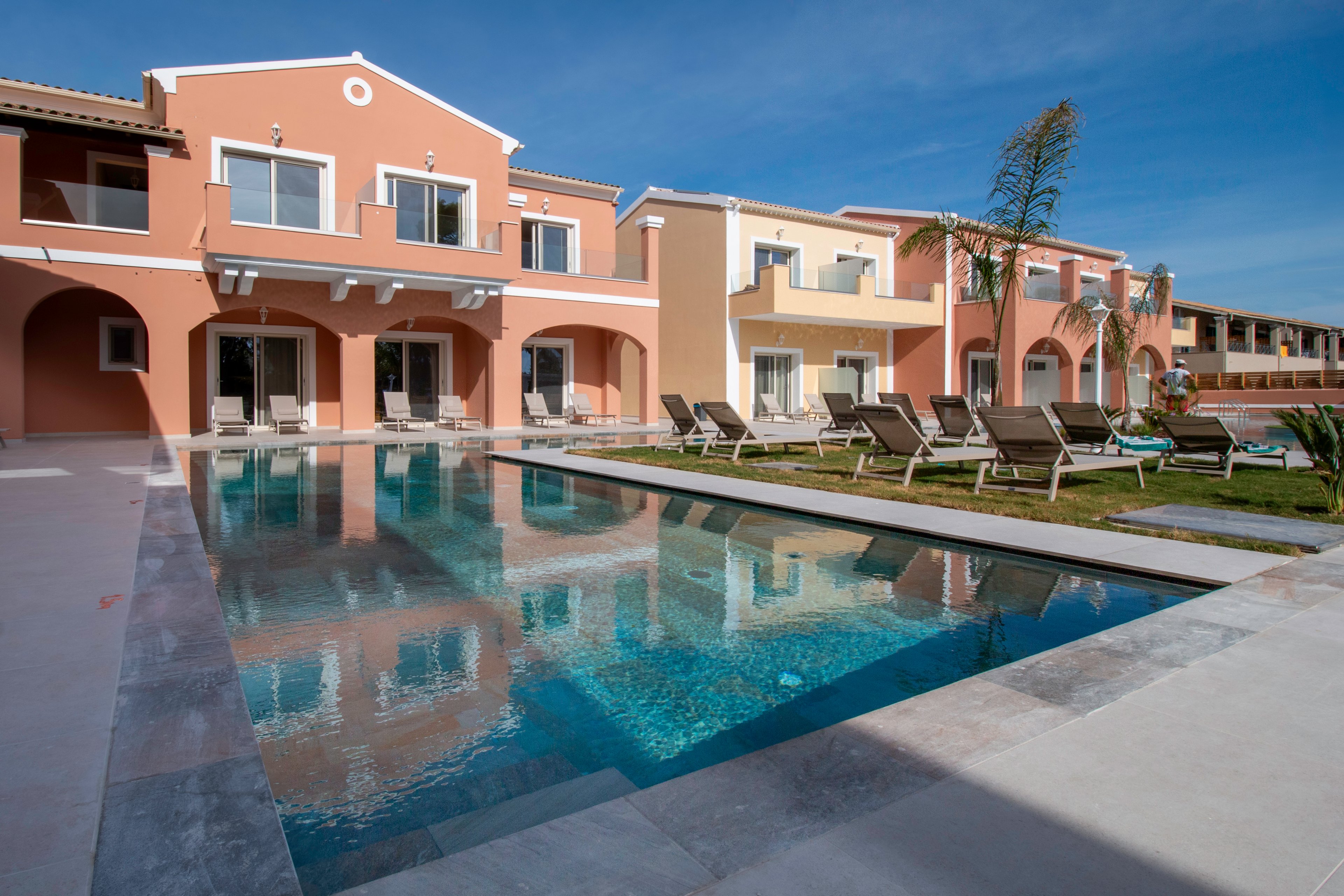 A row of colorful two-story resort buildings surrounds a clear outdoor swimming pool. Sun loungers are arranged neatly along the poolside on a manicured lawn. The architecture features arches, balconies, and large windows reflecting the blue sky. The scene suggests a quiet, sunny vacation setting with no people present.