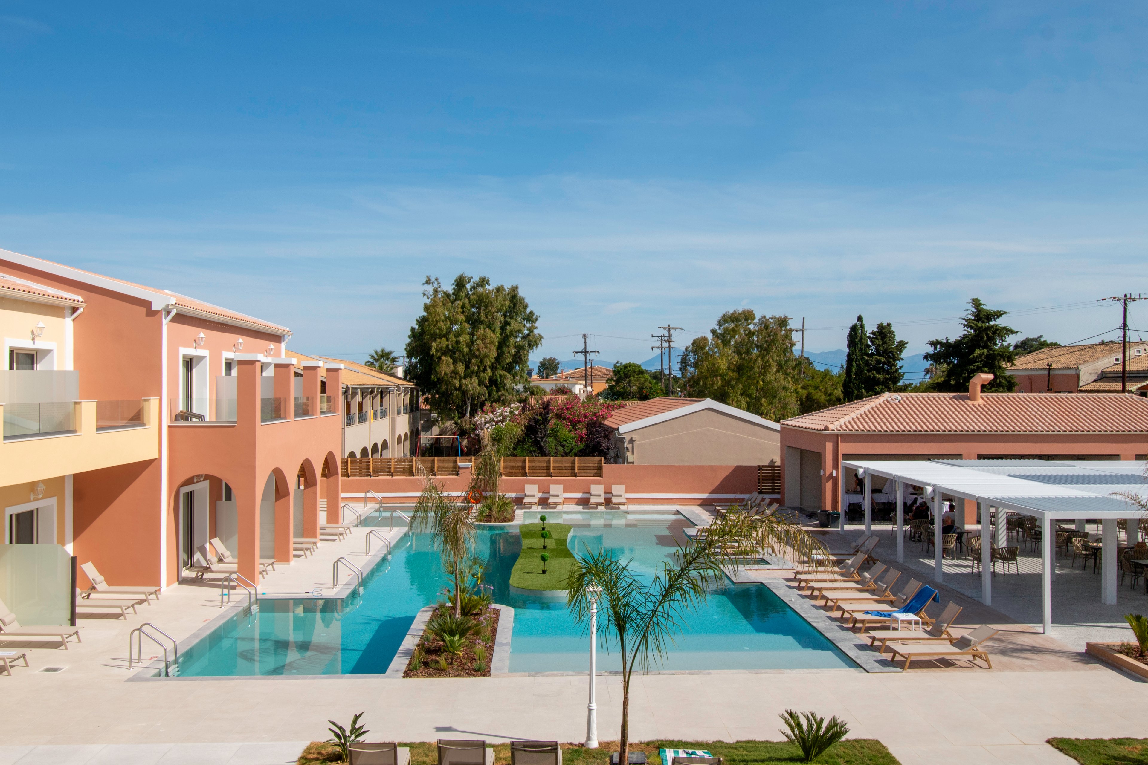 A modern resort courtyard features a long rectangular swimming pool surrounded by peach-colored buildings. Sun loungers and umbrellas line both sides of the pool, creating a relaxing vacation atmosphere. A covered outdoor bar or dining area sits to the right, while small palm trees and greenery add tropical accents. Clear blue sky and distant trees frame the tranquil holiday setting.