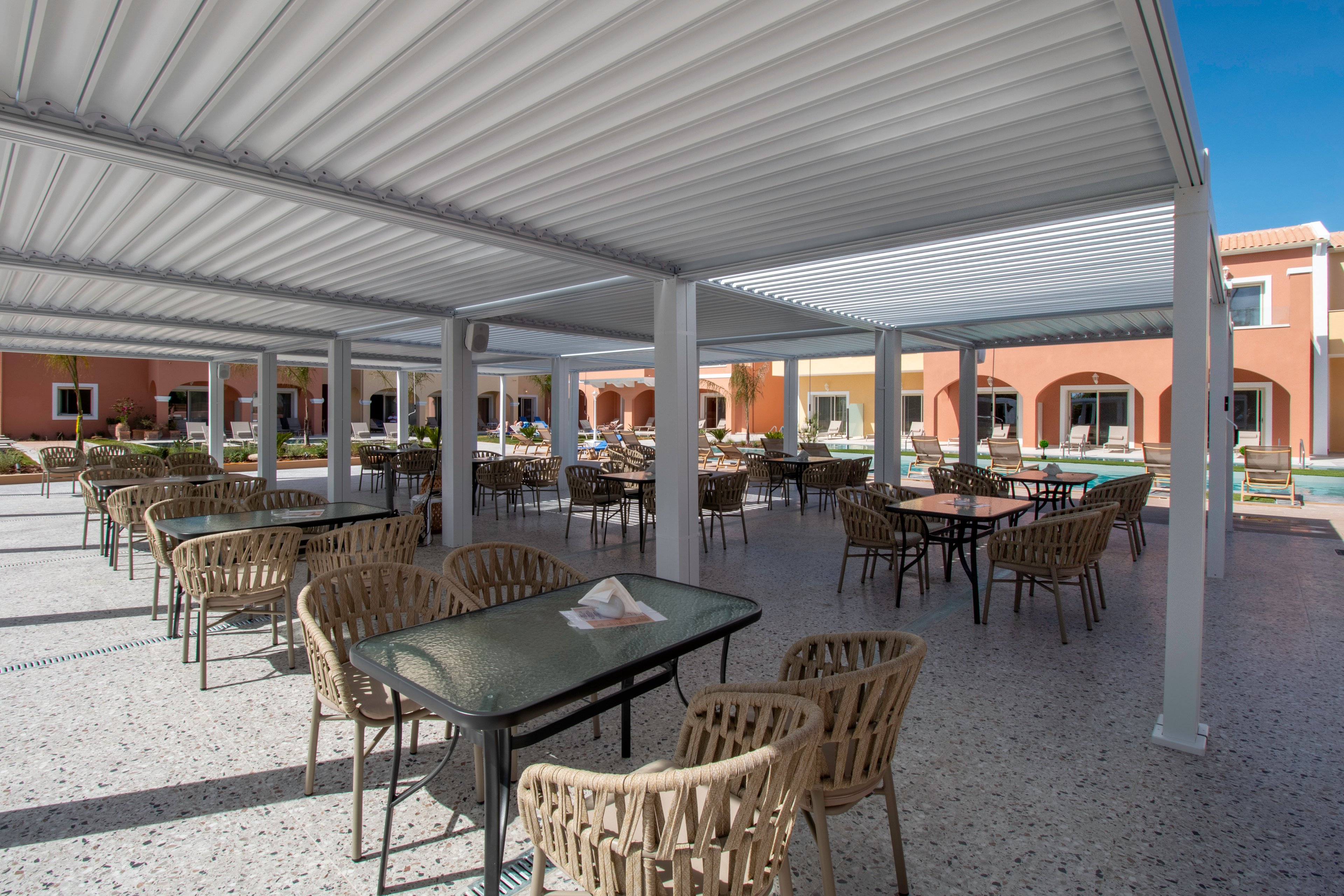 An open-air terrace features multiple tables and woven chairs arranged beneath modern white pergola structures. The setting appears to be part of a resort or hotel courtyard with colorful buildings and a pool area in the background. Bright daylight and clear blue sky create a relaxed, vacation-like atmosphere. The space is empty of people, emphasizing the architecture and ample seating.