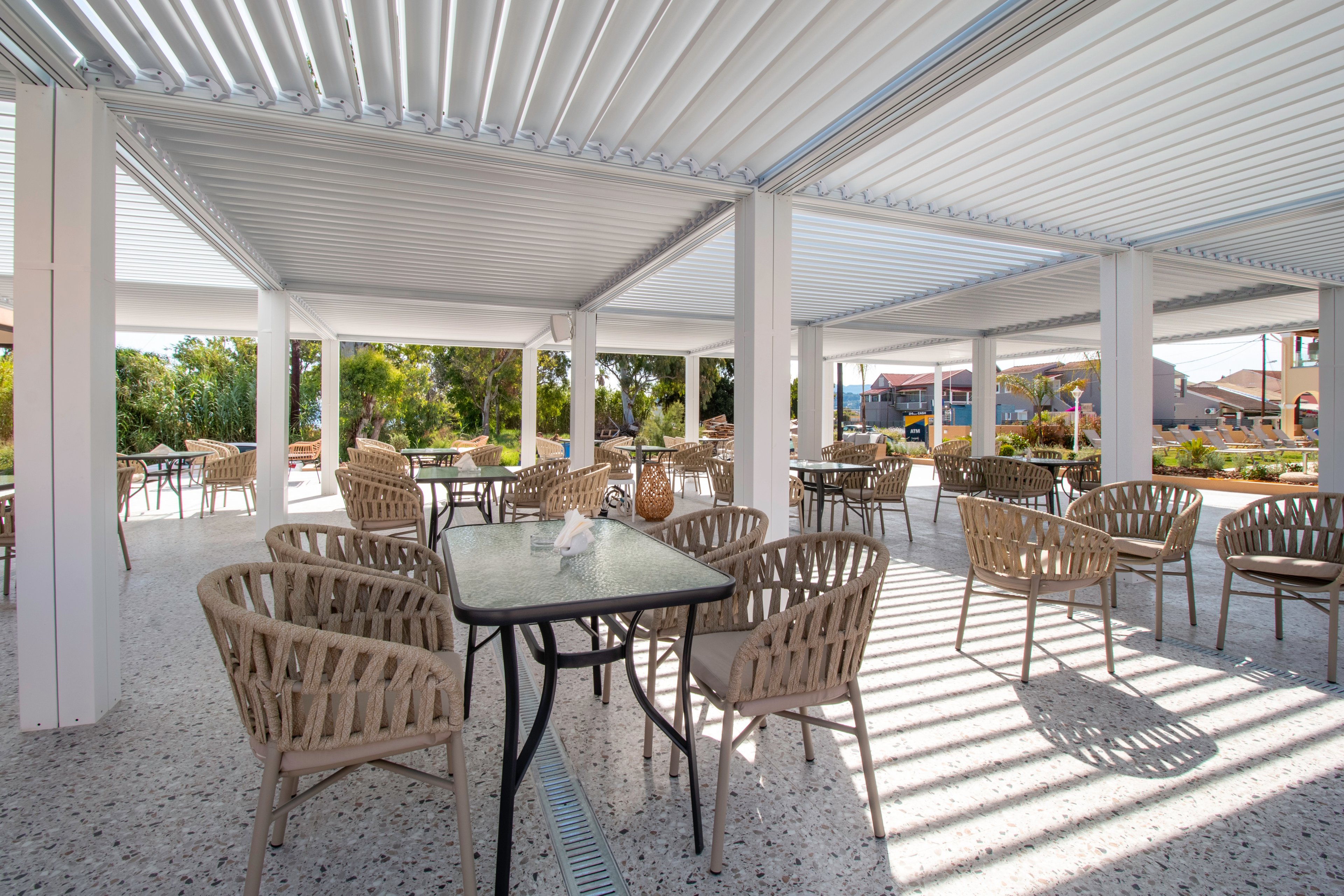 An open-air patio cafe features multiple tables and woven wicker chairs arranged under a modern slatted roof. Sunlight filters through the overhead structure, casting striped shadows on the terrazzo-style floor. The seating area overlooks a landscaped outdoor space with trees and buildings in the background. The scene appears calm and inviting, suitable for casual dining or relaxation.