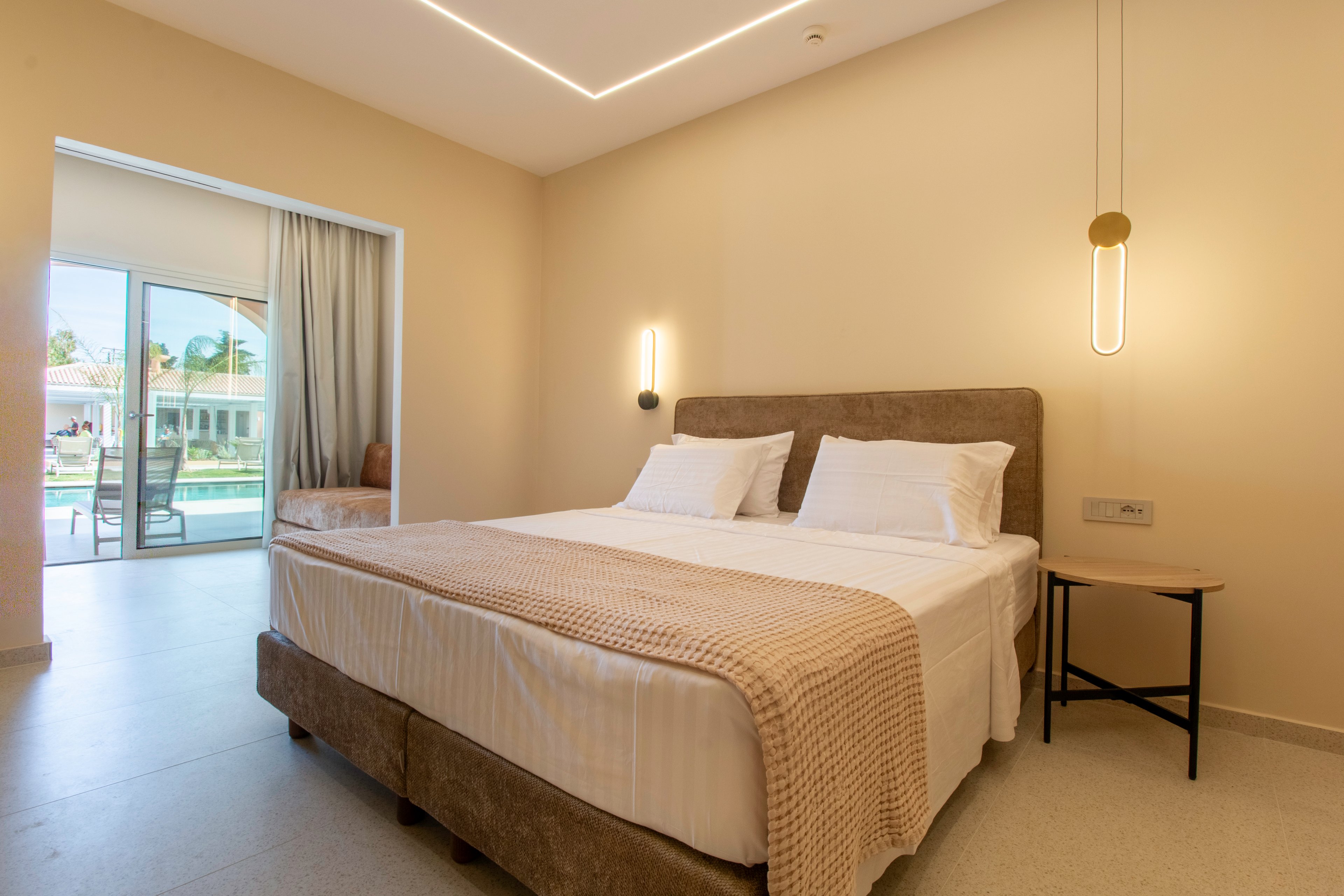 A contemporary hotel bedroom features a large double bed with white linens and a beige textured throw. Warm wall-mounted lights and a minimalist pendant lamp create a cozy ambiance against neutral-colored walls. A sliding glass door opens to a balcony overlooking a pool and resort buildings. The room has a clean, uncluttered design with simple furniture and soft, indirect ceiling lighting.