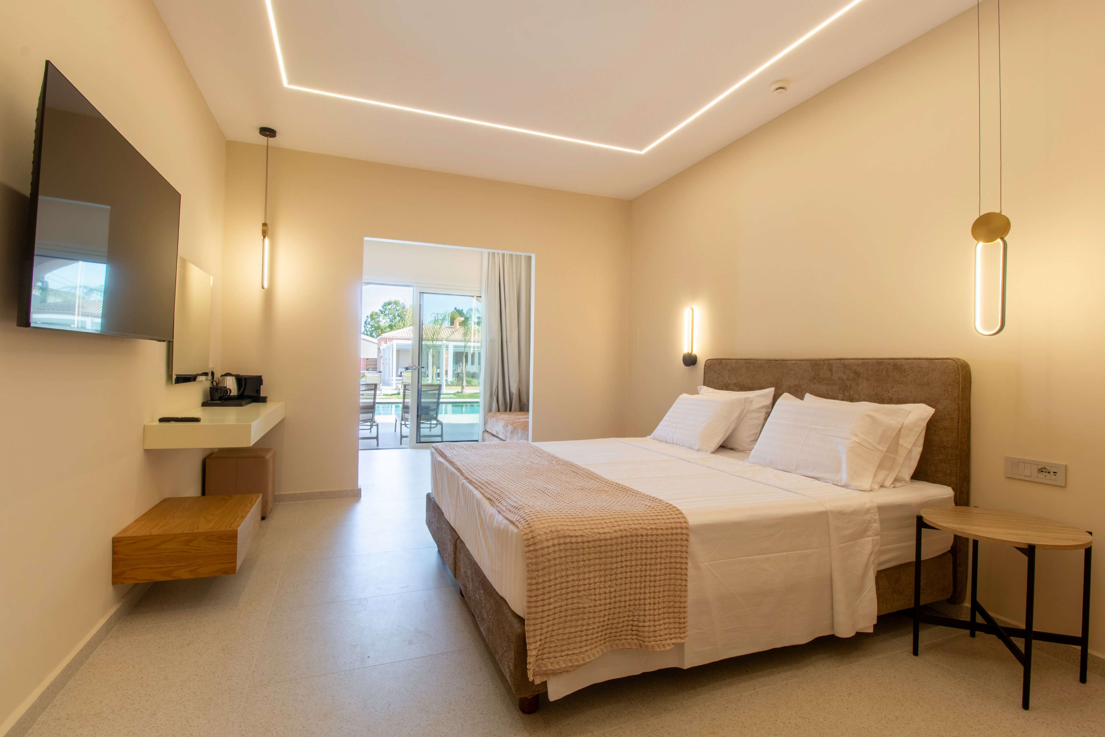 A contemporary hotel bedroom features a large double bed with white linens and a beige throw. Minimalist bedside tables and pendant lights frame the bed, while a wall-mounted television and small desk area sit opposite. Warm neutral tones and indirect ceiling lighting create a calm, inviting atmosphere. Sliding glass doors open to a balcony overlooking a resort-style outdoor area with greenery and buildings.
