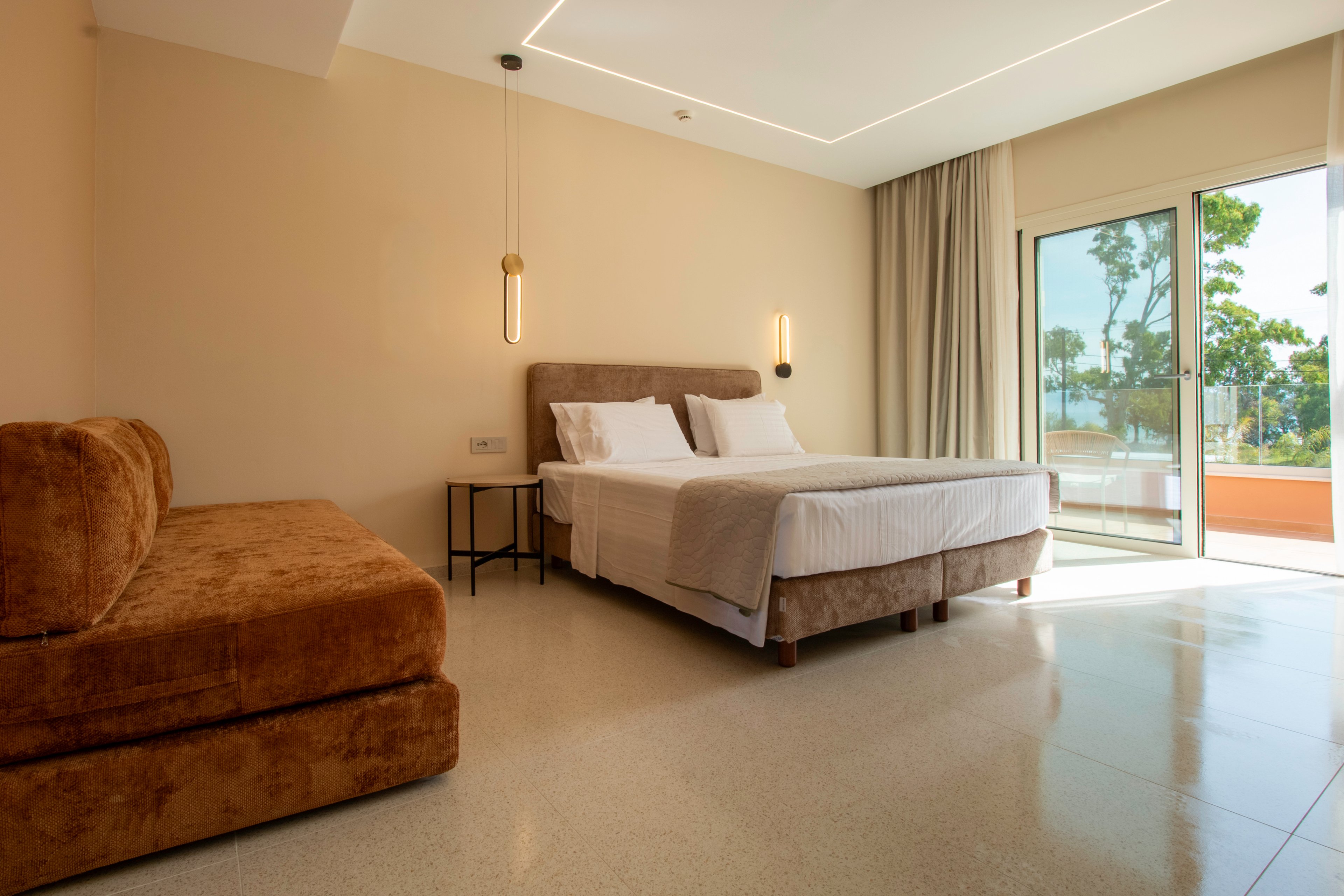 A contemporary hotel bedroom features a large bed with white linens and a matching upholstered headboard. A brown velvet sofa sits along one wall, complementing the neutral beige tones of the room. Minimalist pendant lights and a small side table add modern accents to the clean interior. Floor-to-ceiling curtains frame a glass door that opens to a balcony with trees and a distant water view.