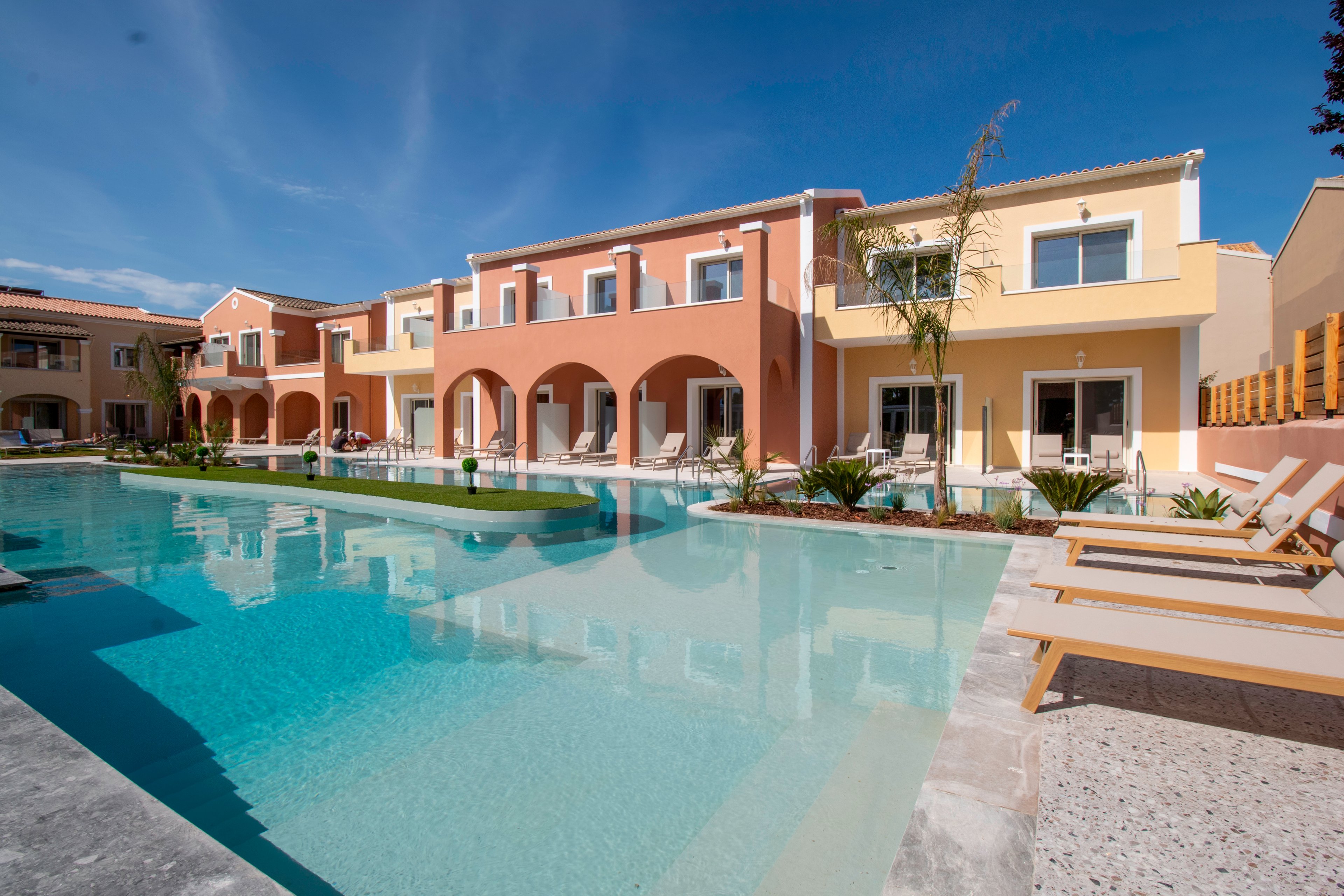 A modern Mediterranean-style resort complex features a large outdoor swimming pool in the foreground. Two-story buildings with arches, balconies, and pastel-colored facades line the poolside. Several sun loungers are arranged along the pool deck, suggesting a relaxing vacation setting. Clear blue skies and calm water reflections enhance the tranquil, sunny atmosphere.